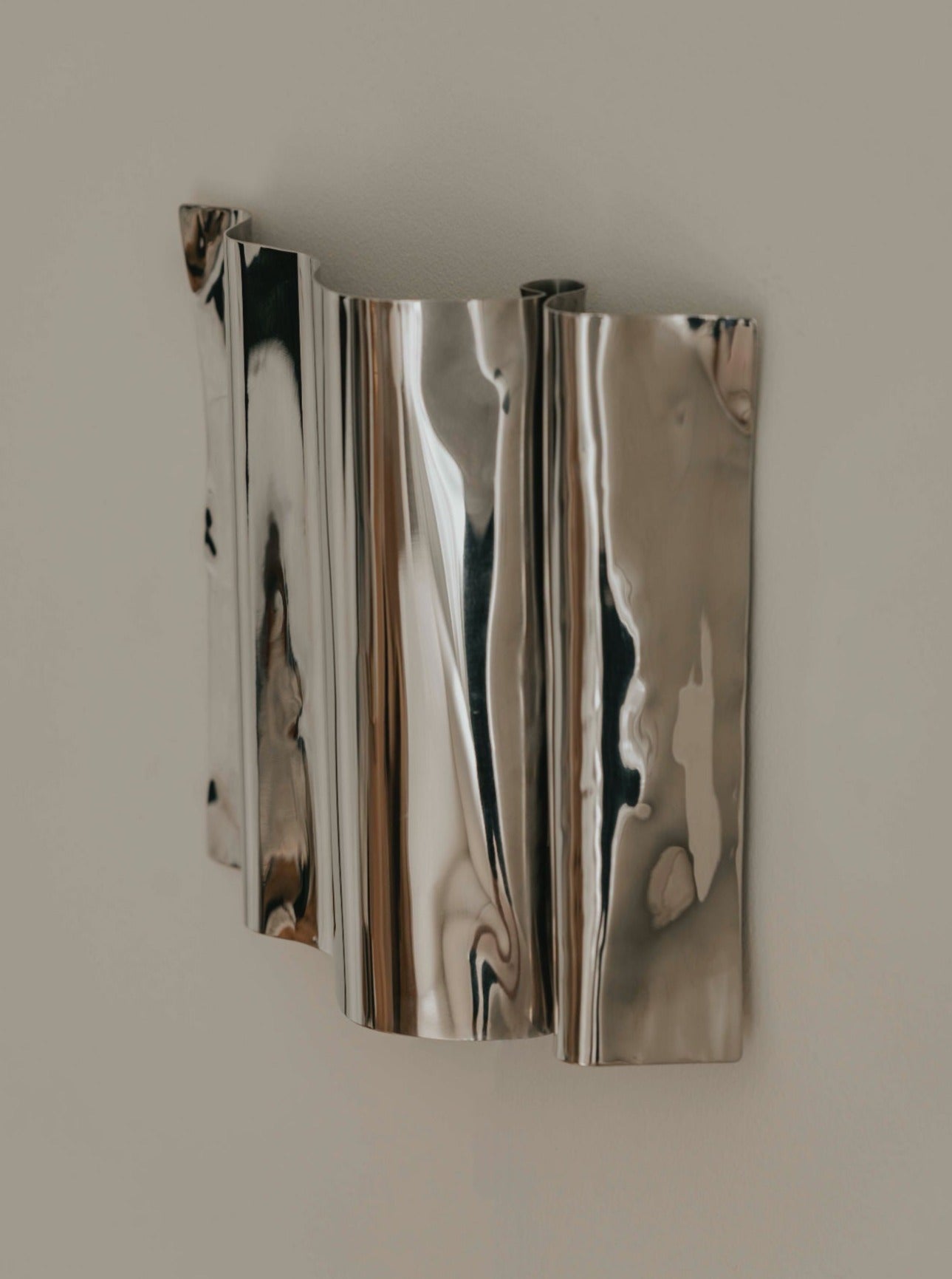 A piece of the Psychedelic Wall Scone by Caia Leifsdotter in polished stainless steel is mounted on a plain wall. The artwork consists of curved, reflective surfaces that distort and reflect light and surroundings, creating unique organic shapes. The metal has a polished, mirror-like finish.