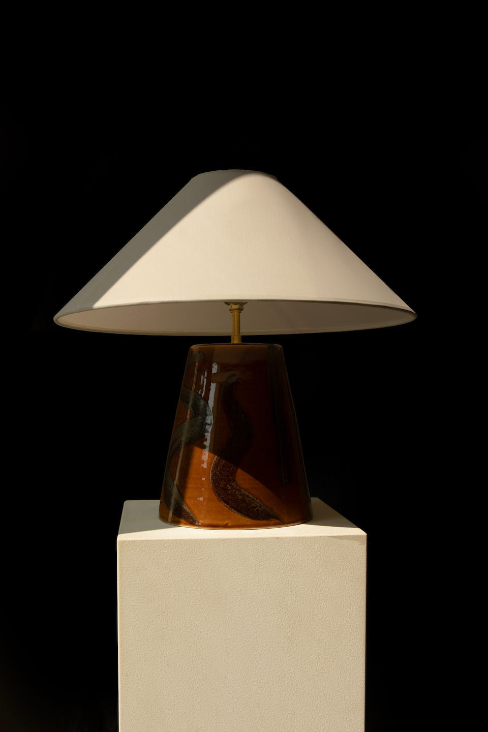 The 'Sierra' Table Lamp by Landy Rakoto Ceramics features an elegant conic shade atop a white pedestal. Its glossy, dark, textured base is adorned with an amber glaze, all set against a solid black background enhancing its striking design.