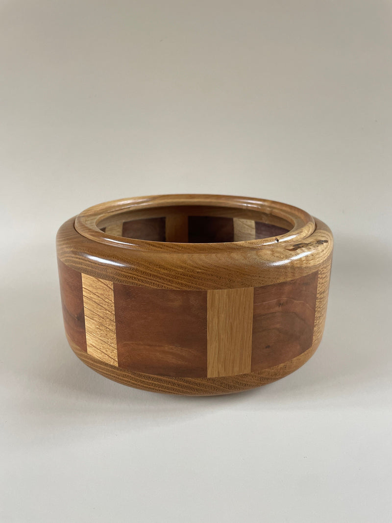 Introducing the Hand Turned Lidded Circular Box by Maud Vaughan. This exquisite box is skillfully crafted from wood and features a seamless, polished appearance with an alternating pattern of oak, walnut, and chestnut tones. Its design boasts a wide rim and a smooth finish that gracefully contrasts against a plain, light-colored surface.