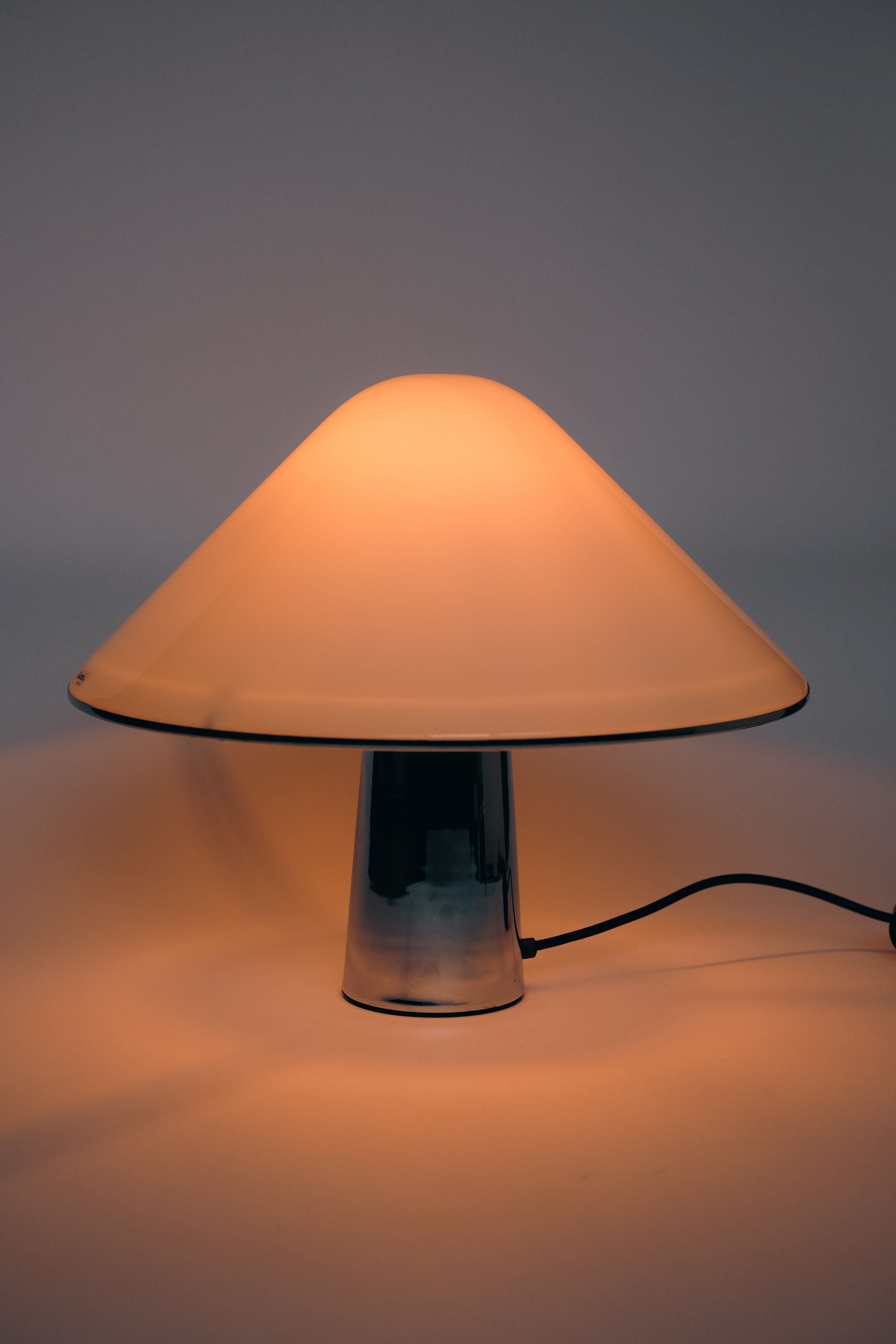 The Harvey Guzzini Elpis Table Lamp by septembre studios, inspired by Luigi Massoni, features a mushroom-shaped metallic shade for a warm glow. Its sleek reflective base and black power cord enhance its vintage appeal against a neutral backdrop.