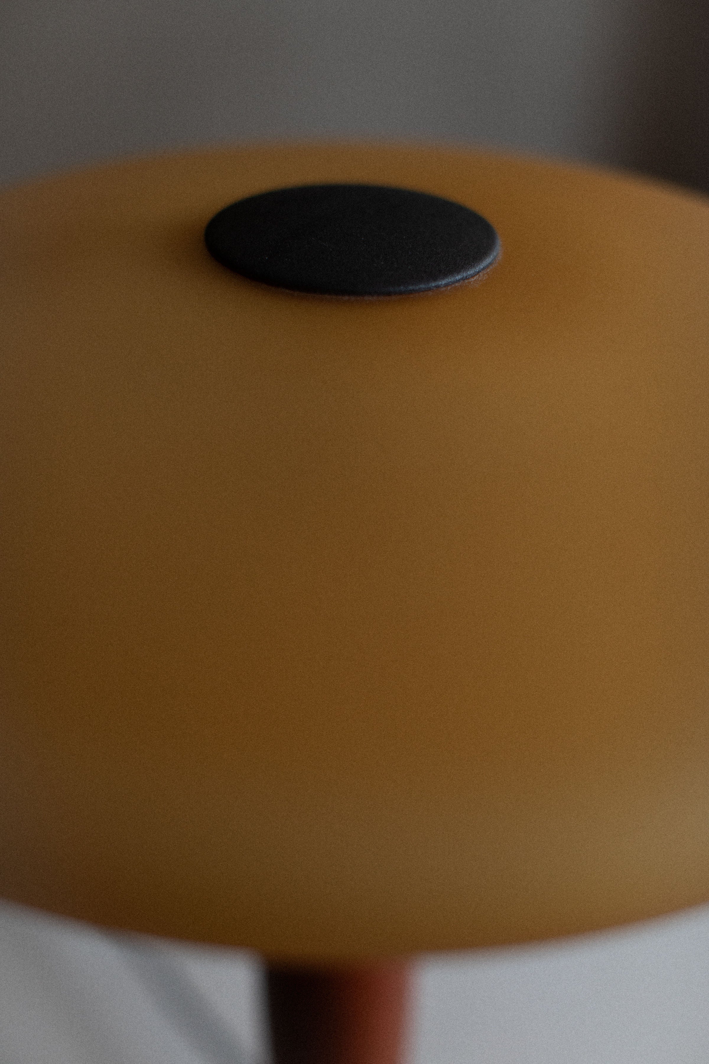 Close-up of a brown stool with a black round cushion centered on the top, set against a softly blurred background featuring an Out For Lunch vintage Murano lamp.