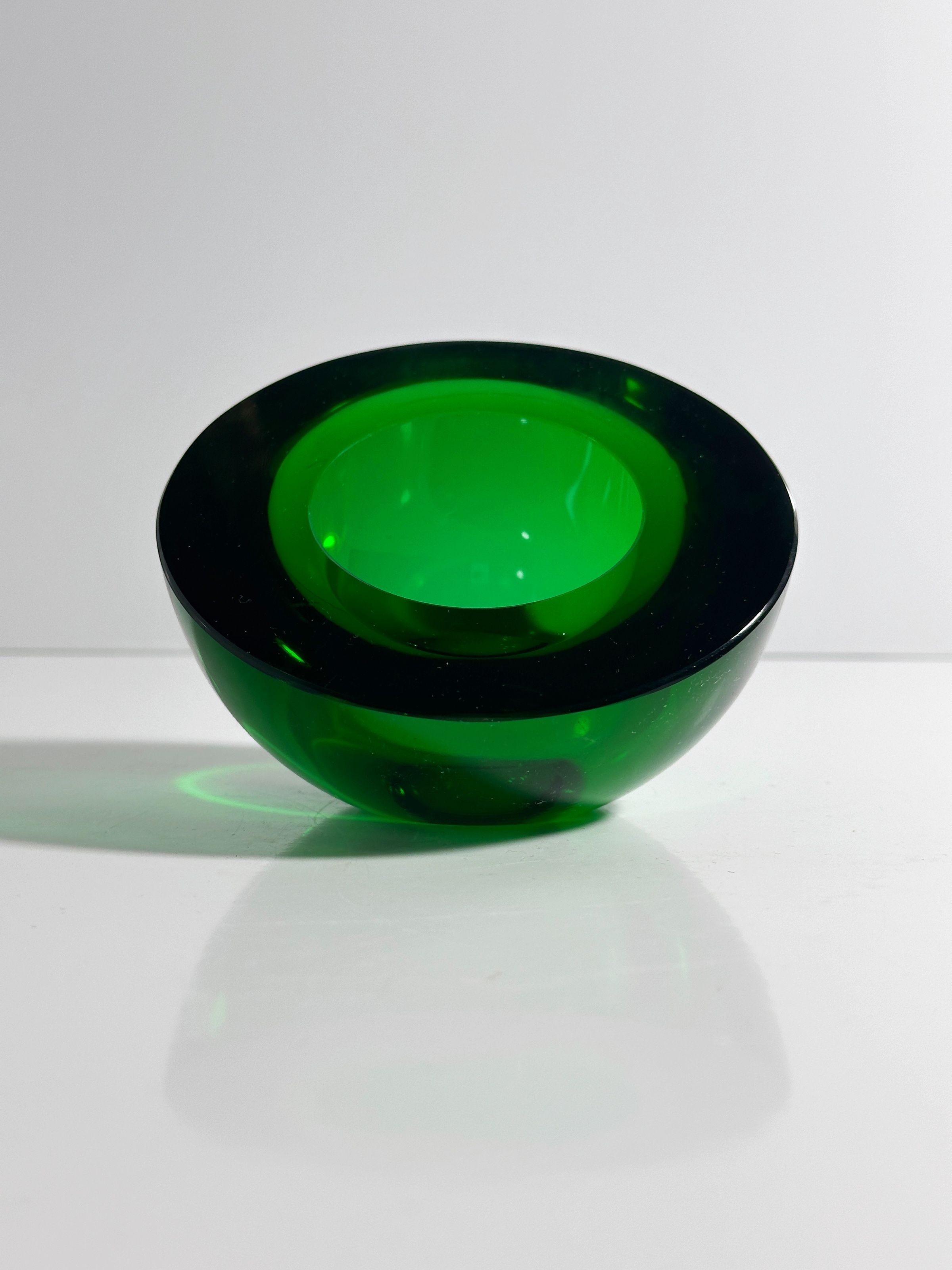 A glossy, emerald green "1960s Space Age Glass Ashtray" by Dodo Vintage rests on a white surface. The light reveals its translucent, smooth, curved design against the minimalist background, echoing 1960s artistry.