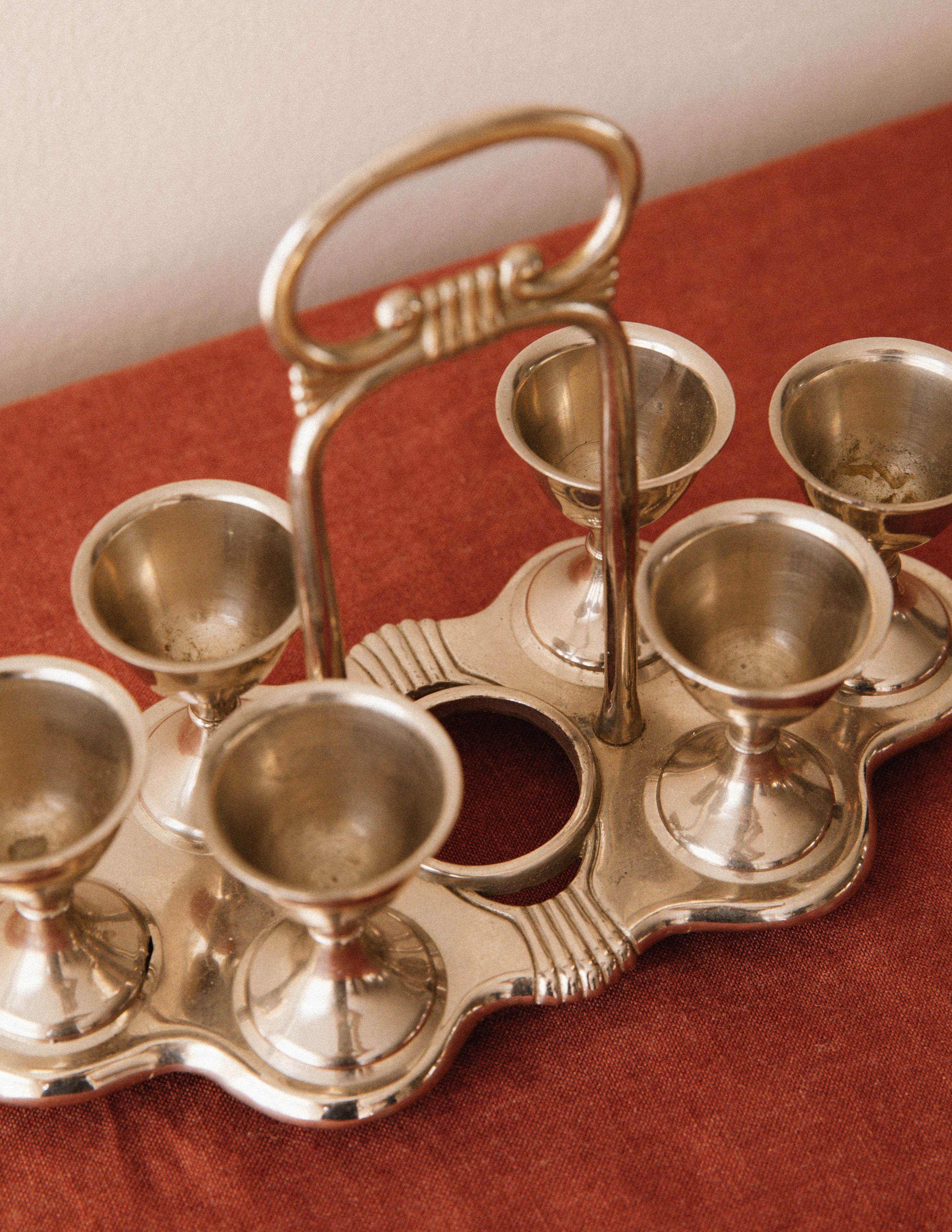 The Arcā Collections Vintage Egg Cruet Holder Set features a central handle and ornate detailing, with six sculpted cups, making it a striking tabletop accent. It rests on a reddish-brown surface.