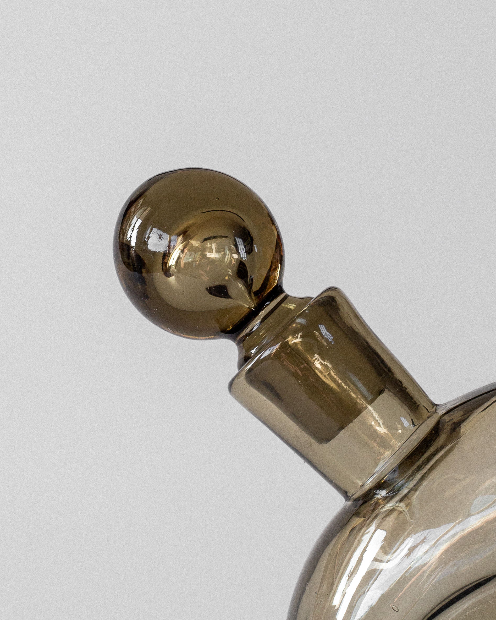 The Glass Decanter by Harald Notini from the 1930s, designed by Spigel, features a smoky brown glass with a decorative round stopper against a plain gray background. This modernist piece reflects the elegance of Swedish modernism.