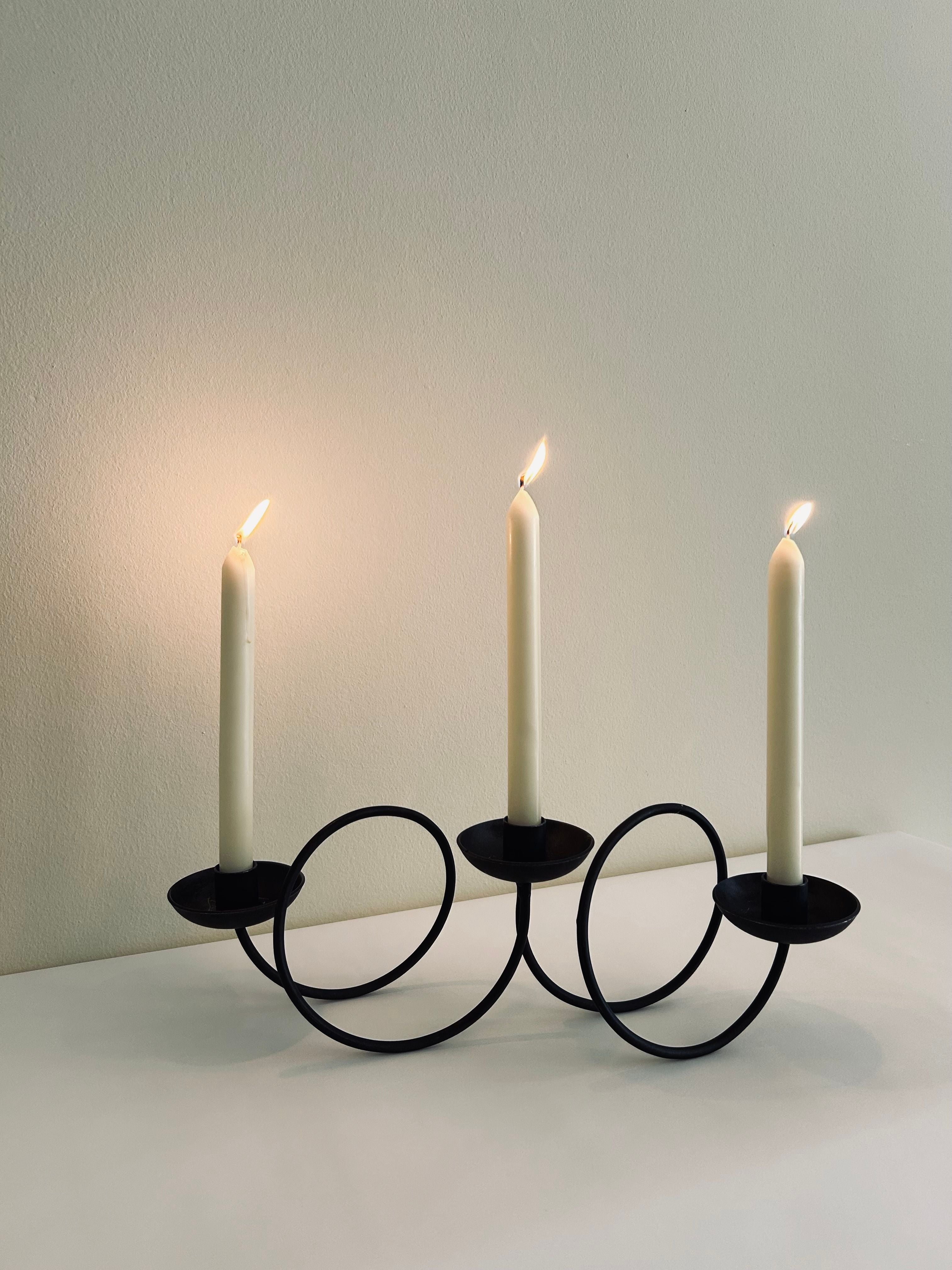 The Brutalist Swirl Candle Holder by Flock Studio, a modern black metal piece with a geometric loop design, stands elegantly on a light-colored surface against a beige wall. It features three lit white taper candles that beautifully enhance its unique aesthetics.