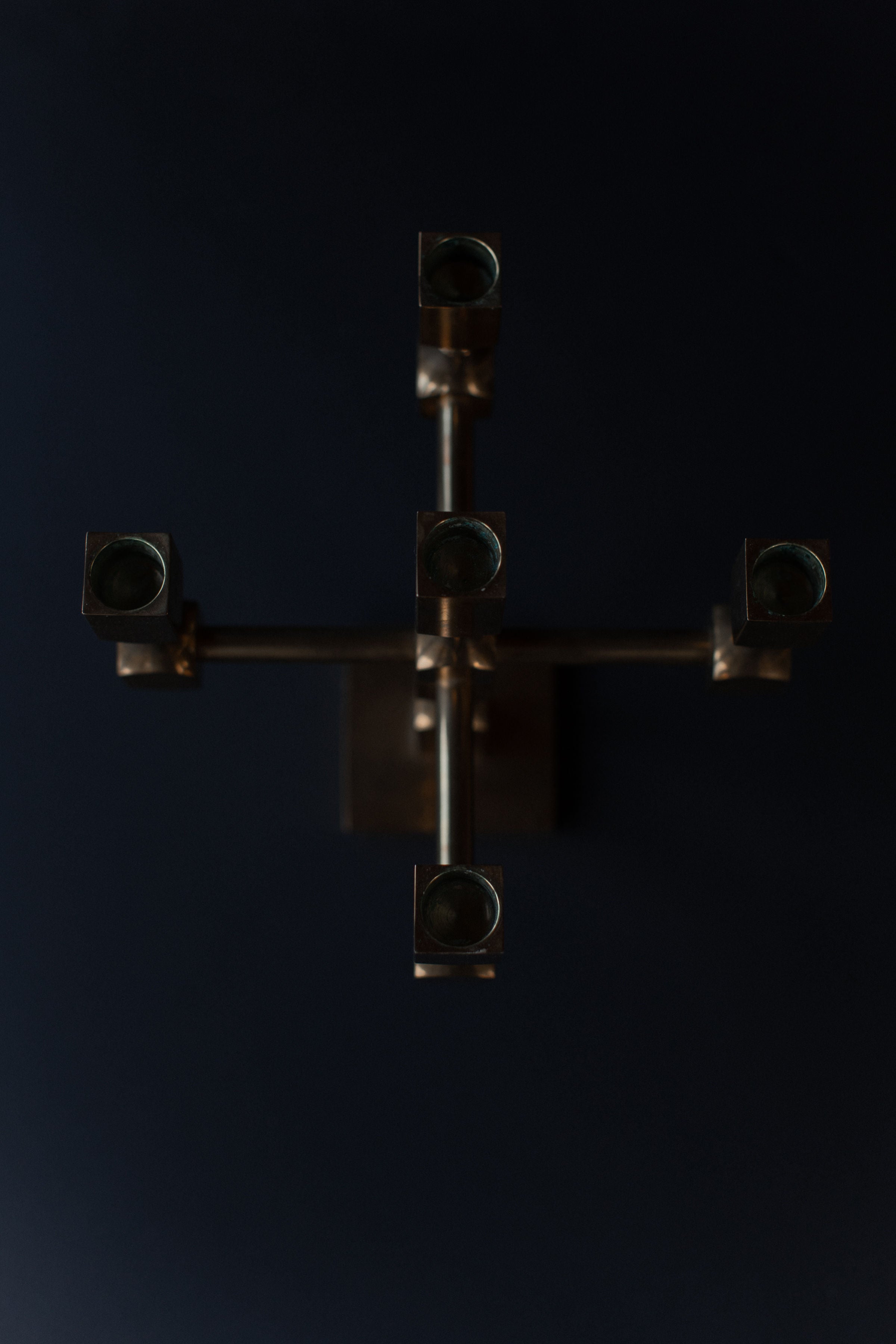 The Brutalist Brass Candleholder by Out For Lunch is an overhead view of a modern, industrial wall-mounted piece with six cylindrical solid brass holders on metallic arms against a dark background.