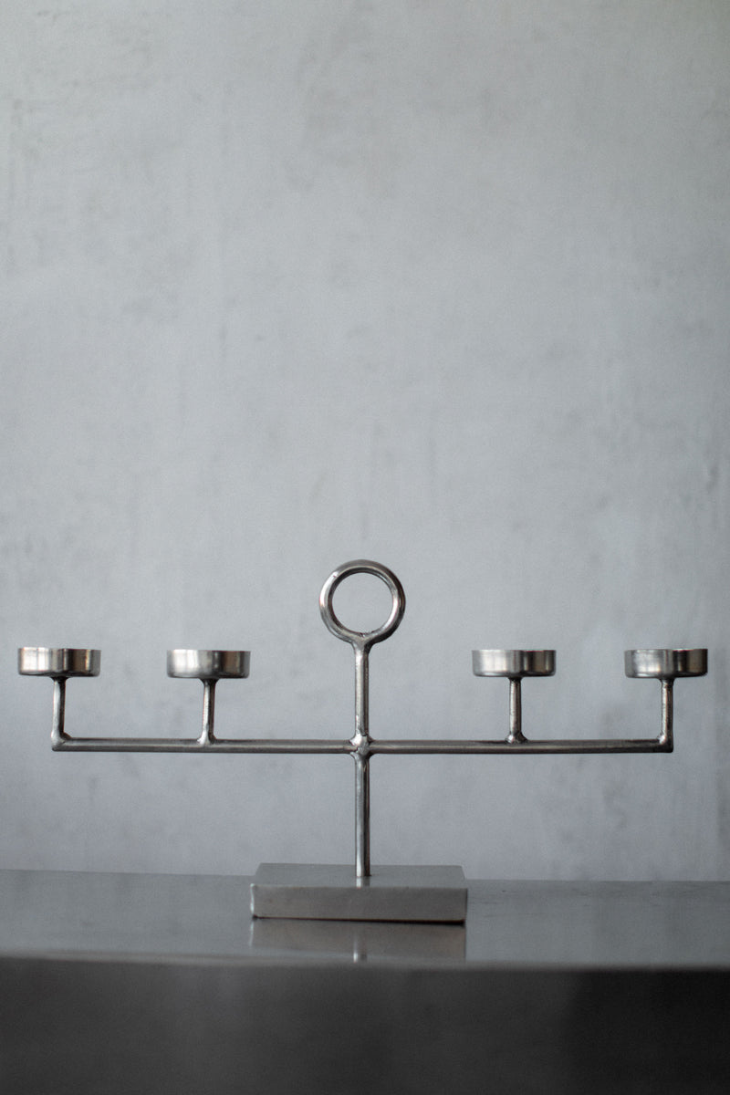 The Out For Lunch Steel Candleholder showcases a minimalist, geometric design with five symmetrically arranged holders and a central circular handle, elegantly resting on a smooth, dark surface against a light gray background.