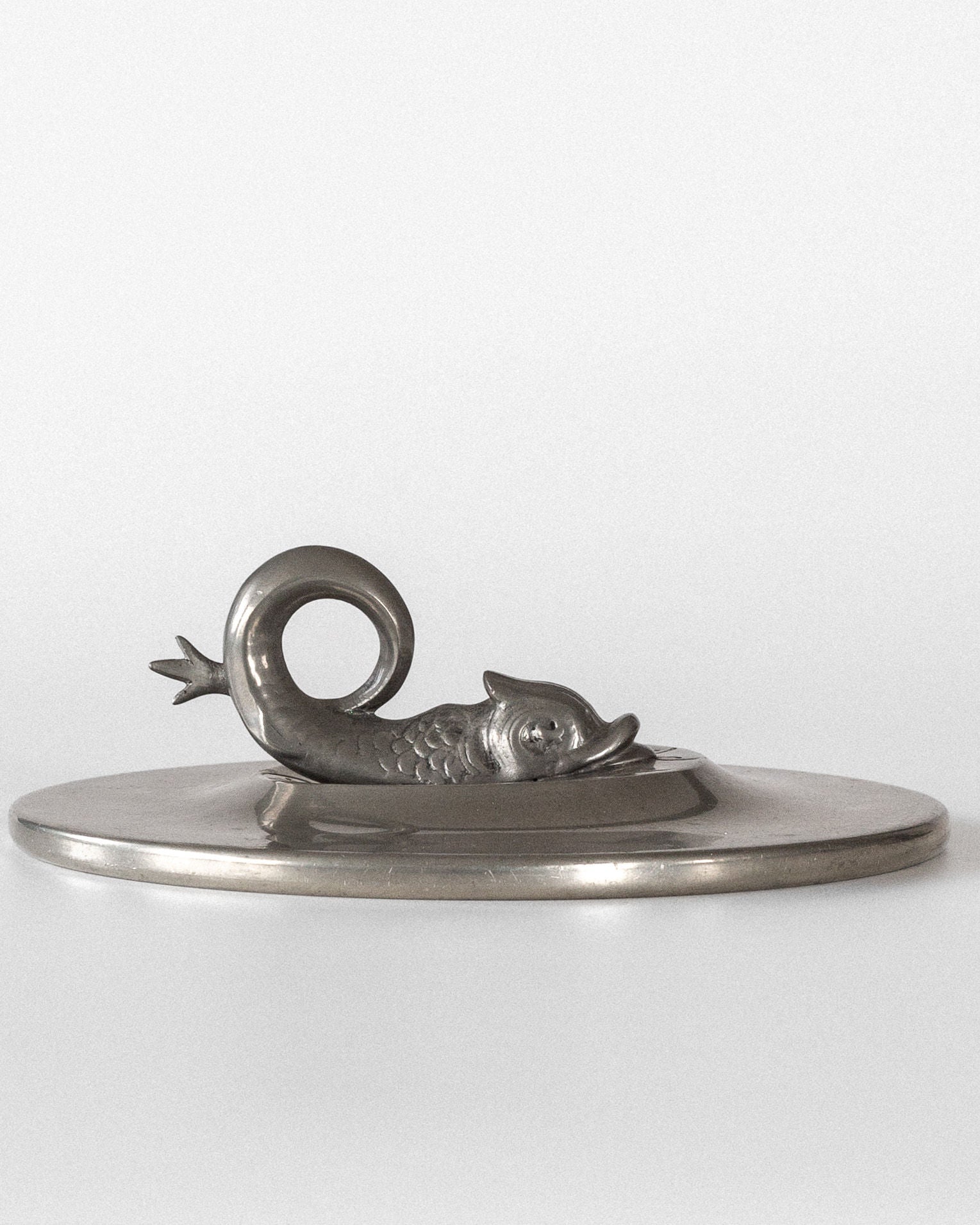 The Spigel Art Deco Metal Mirror with Koi Fish, from the 1920s, features a pewter lid with a handle shaped like a curled fish or dolphin. Intricate scale detailing and a curved tail capture the essence of Swedish Grace against a plain, light background reminiscent of Art Deco elegance.