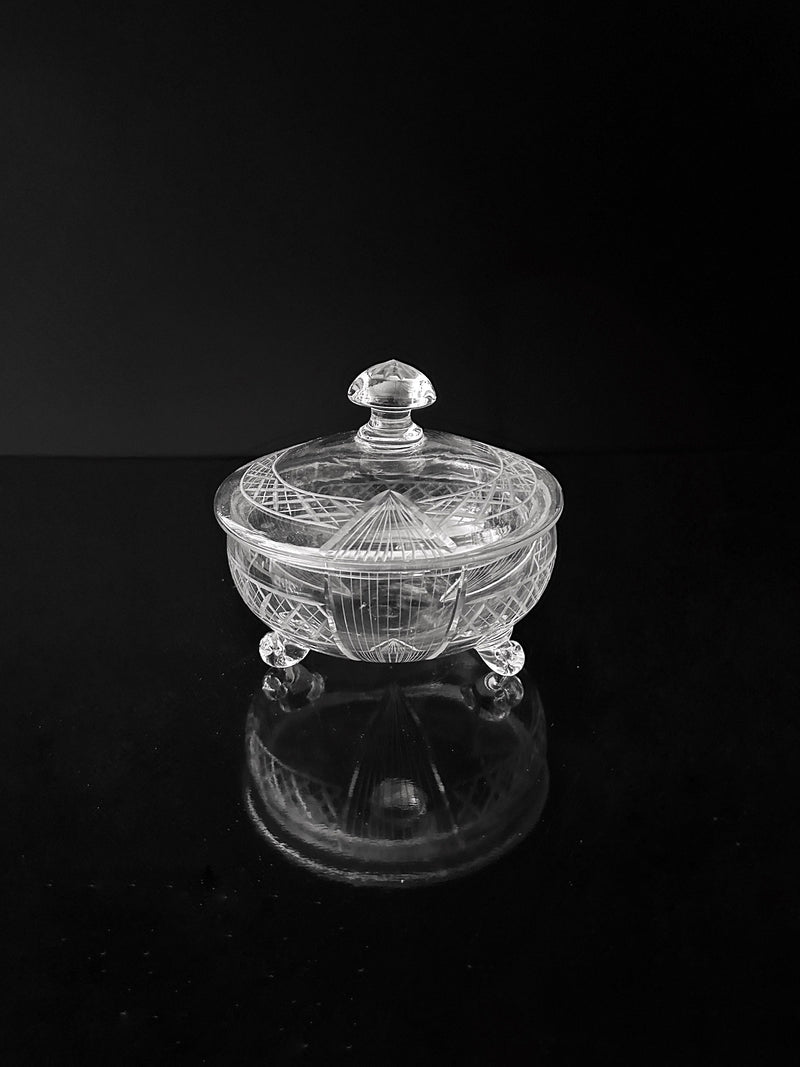 The Art Deco Glass Bonbonnière - 1930 from Dodo Vintage, displaying an intricate Art Deco pattern, sits on a reflective black surface. This elegant dish, complete with a matching lid featuring a knob handle, gracefully rests on three small curved feet.
