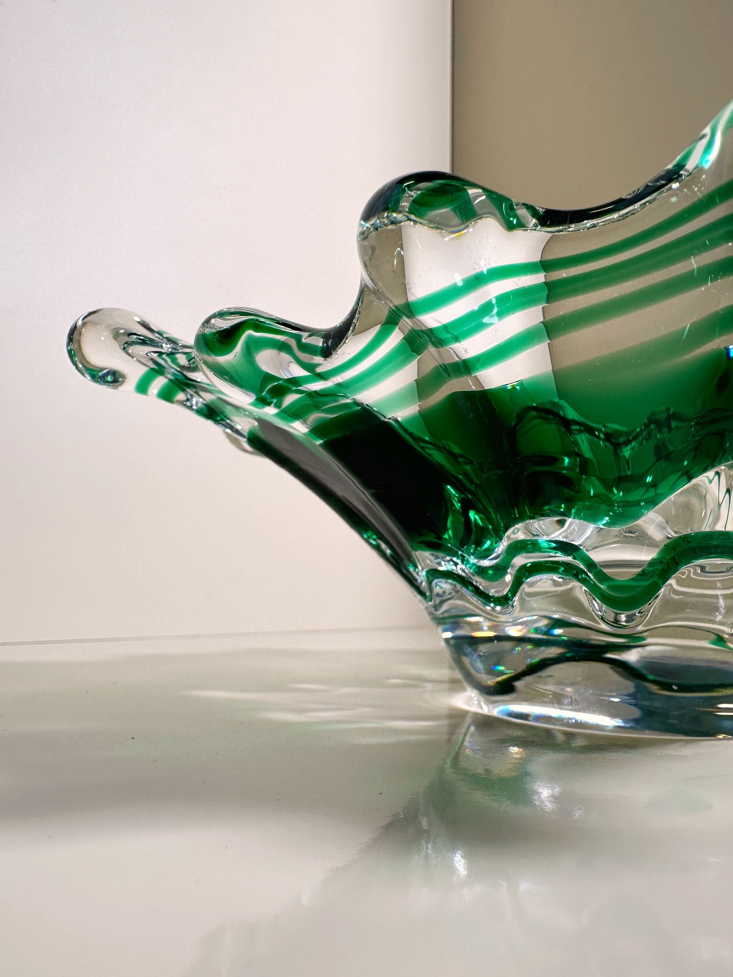 A close-up photo of the Dodo Vintage Green Swirled Murano Vide-Poche shows intricate green and clear stripes with wavy edges casting shadows and reflections on a white surface. The softly lit background highlights the bowl's elegant craftsmanship and transparent glass design.