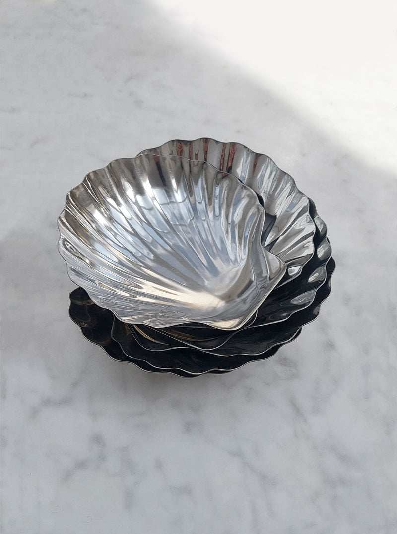 A stack of shiny, fluted metal bowls of varying sizes is placed on a light gray marble surface. Resembling the Set of 6 Small Shell Dishes by Les Objoies, the bowls have a scalloped design and appear slightly reflective. The top bowl is small and rests inside the larger ones beneath.