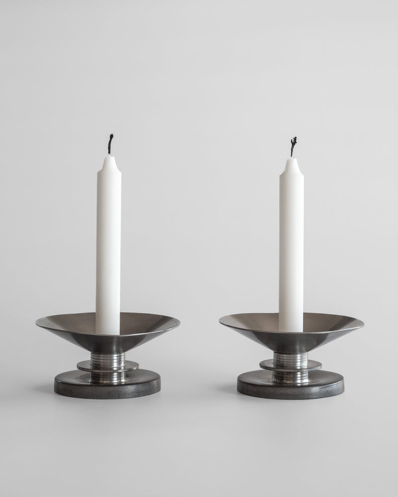 Two white candles on Modernist Candleholders by Sylvia Stave 1930s from Spigel stand against a plain gray background. These 20th-century pieces feature a wide, flat base with a cylindrical center, supporting the unlit candles. The blackened wicks suggest previous use, adding character to the design.