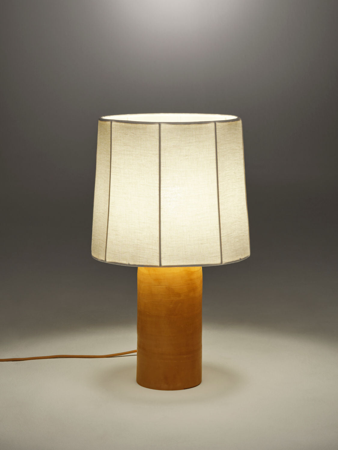 A softly lit Sonora Linen Table Lamp by Nassi Lamps with a cylindrical, terracotta base and a large, cream-colored, pleated fabric lampshade. The lamp is placed on a plain surface against a gradient gray background, with its power cord visible trailing on the left side.