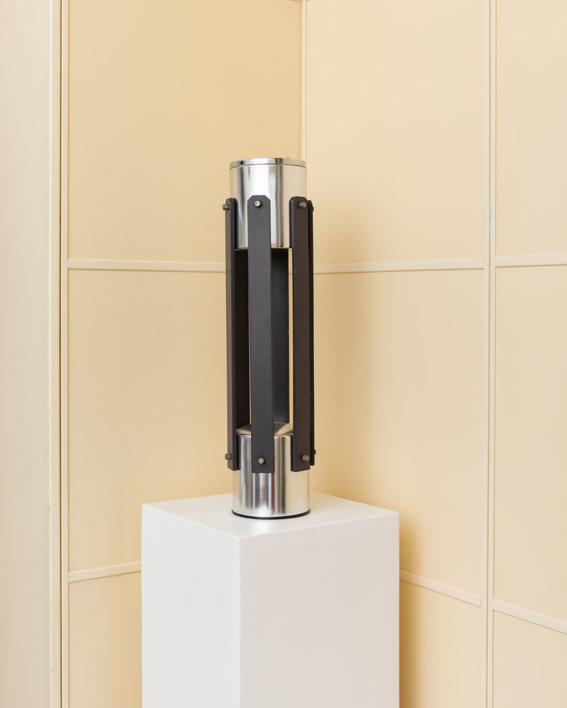 The Metal Floor Ashtray by Bottega Jacobs, Italy 1970s, features sleek vertical black panels on a cylindrical metal form atop a white pedestal, recalling modern space age design against a beige tiled wall.
