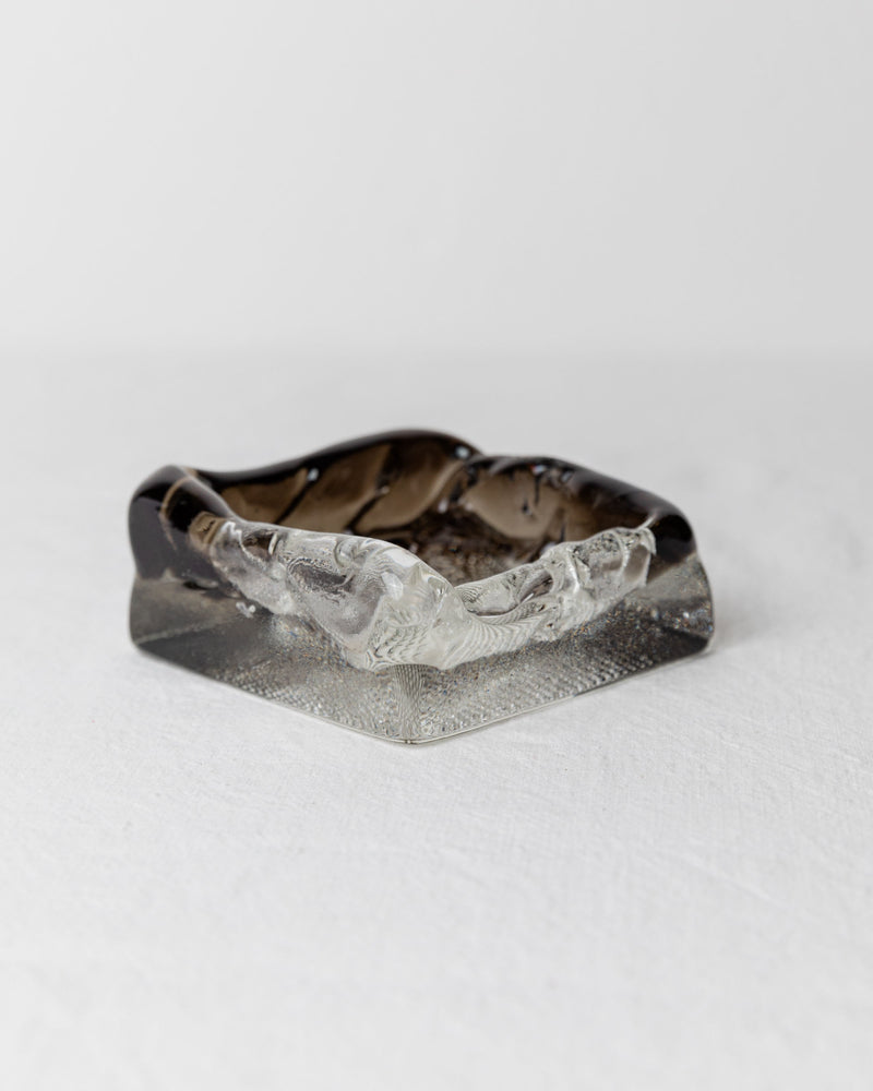 On a plain white surface sits the Vintage Murano Glass Ashtray from the 1980s, crafted by Bottega Jacobs. This square, dark gray ashtray features a wavy, textured rim that combines transparent and smoky glass. Its design encapsulates the Hollywood Regency style, offering a modern yet vintage charm reminiscent of classic Murano craftsmanship.