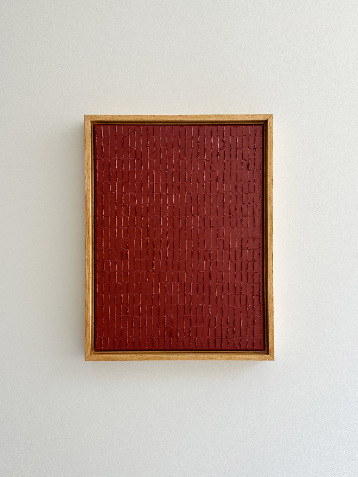 A piece of textured red artwork, "Indian Red - Oil on canvas" by Roi Elmaliah, with a pattern resembling vertical brickwork and inspired by Korean Dansaekhwa, is displayed in a simple wooden frame against a plain white wall.