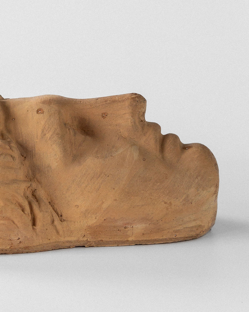 A Terracotta Wall Mask Planter by Spigel is depicted on its side against a neutral backdrop. The piece showcases smooth contours, a closed eye, and a serene expression, highlighting the natural texture of terracotta.