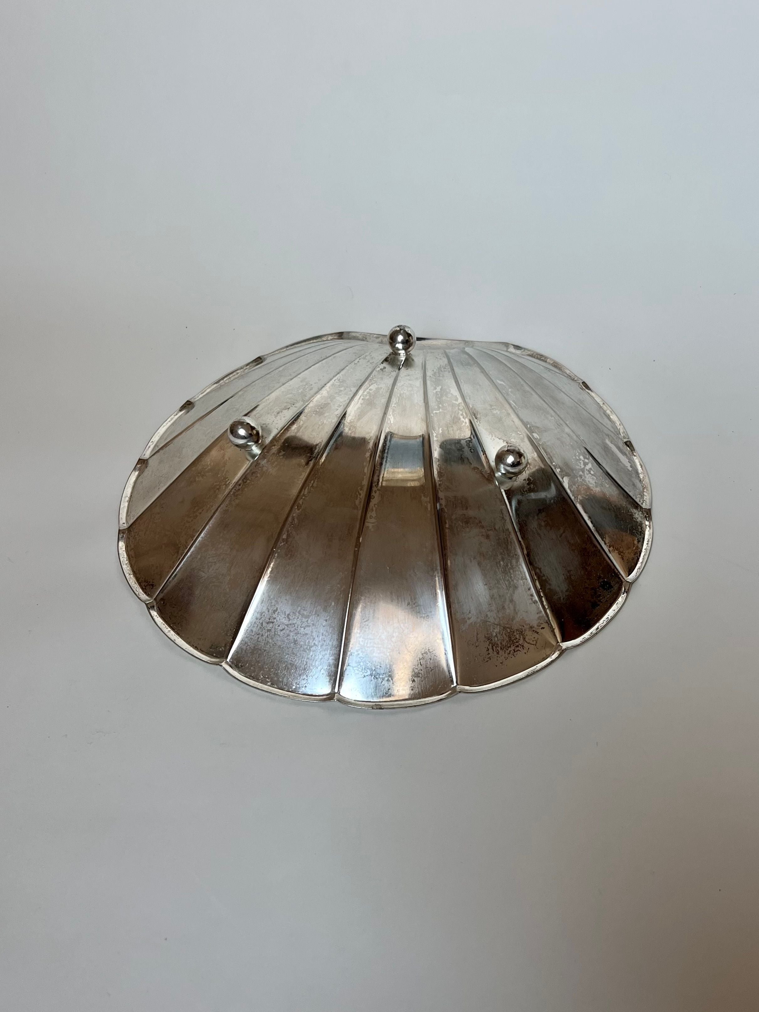 A round metallic object with a dome-like design reminiscent of Manushi's Grand Silver Shell Dish, sourced in France. It has a shiny, reflective surface and three small, evenly spaced spherical knobs on top, set against a plain, light-colored background.