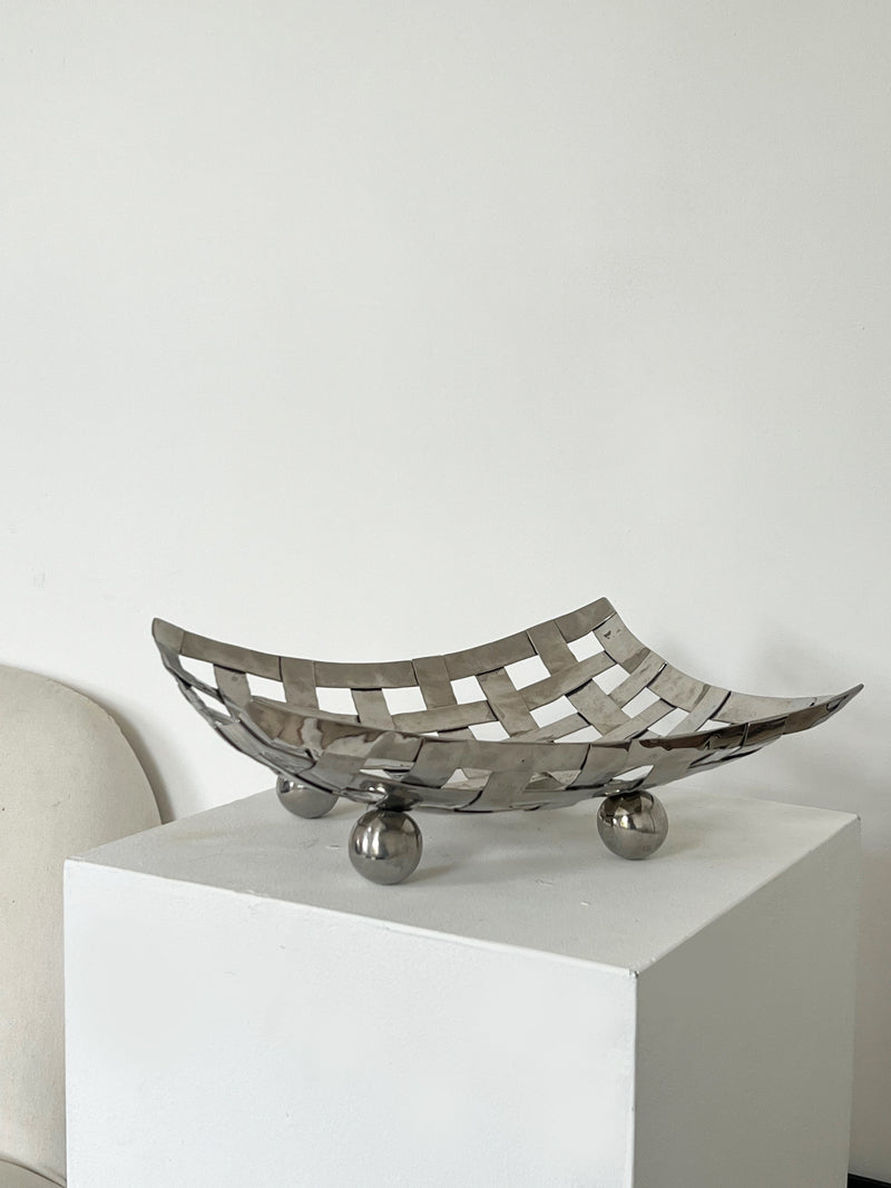 The Porter Studio's Aluminum Dish on Feet, a metallic woven basket with elegant proportions, rests on a white pedestal. Its grid-like pattern is elevated by three ball feet.