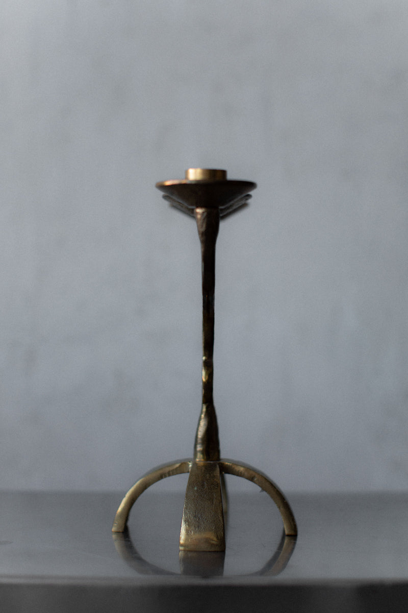 A Brutalist Brass Candleholder by Out For Lunch, showcased on a light gray background, highlights artisanal craftsmanship. It features a simple, slender design with a wide base, capturing a vintage aesthetic.