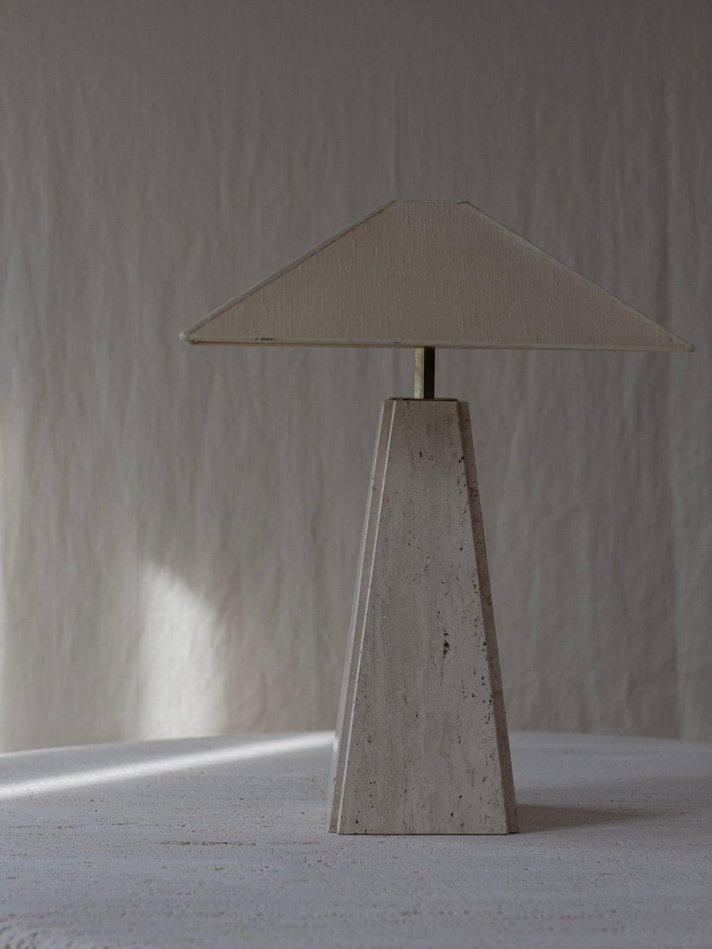 This Camille Breesch-inspired Travertine Table Lamp by Bicci de' Medici features a minimalist design with a beige triangular shade and rectangular stone base. It is elegantly placed on a textured white surface against a plain, light-colored wall casting subtle shadows.