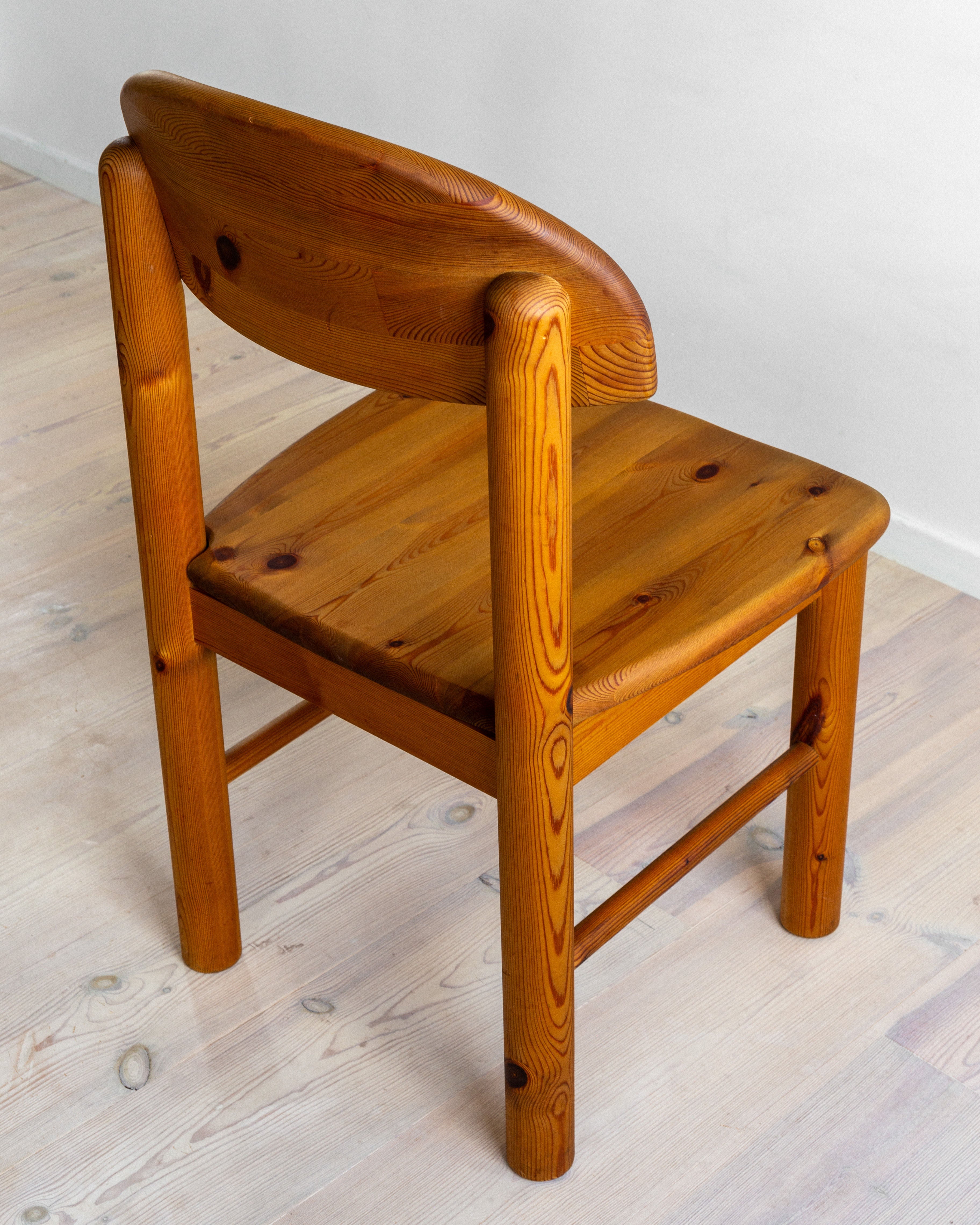 A set of four Pine Chairs by Rainer Daumiller from the 1970s, under the Spigel brand, is gracefully arranged at an angle on a light-colored wooden floor. These chairs with curved backrests and smooth polished finishes exemplify the solid strength of pine furniture, featuring sturdy legs, reinforced seats, and timeless Danish design elements.