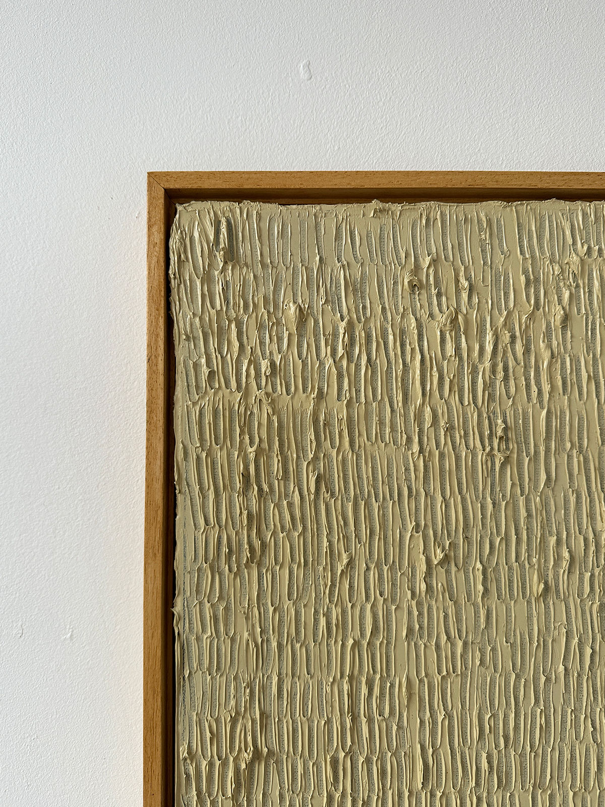 The "Buff Titanium" painting by Roi Elmaliah features textured beige vertical ridges filling a wooden-framed canvas, crafted using oil on canvas. The artwork showcases a repetitive pattern of raised brush strokes on a neutral background, creating a tactile, minimalist aesthetic. This piece is displayed on a plain white wall in Amsterdam, The Netherlands.