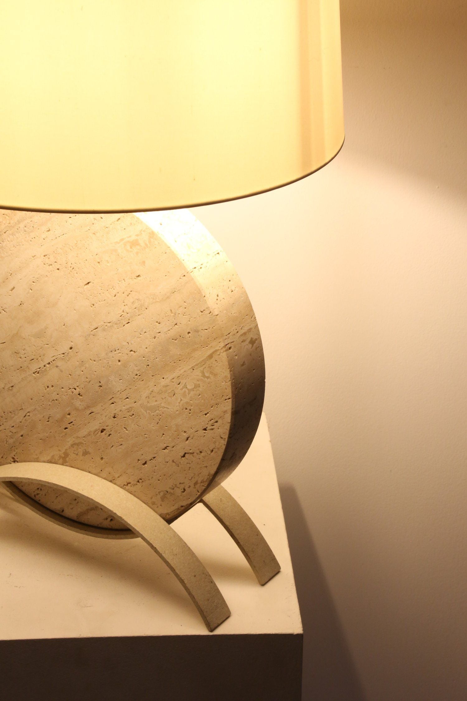 The Italian Travertine Table Lamp by Ancien et Jolie features a beige cylindrical shade and sits on a light surface. Its sculptural design includes a round, textured base with curved supports and clean lines, casting a warm glow onto the wall behind it.