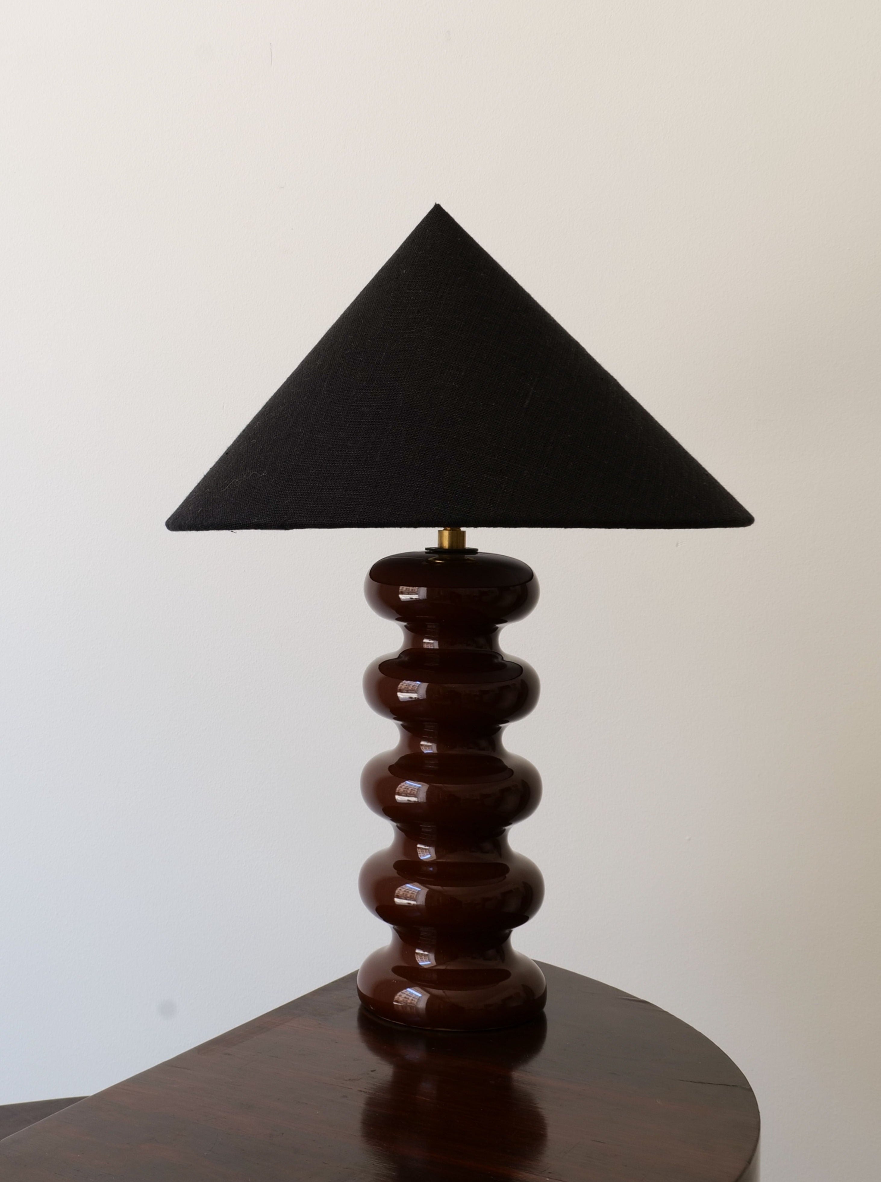 A modern lamp with a handmade, black, pyramid-shaped lampshade and a glossy, tiered, brown ceramic base. The Sculptural Ceramic Lamp by Collection Apart is placed on a corner table against a plain white wall.