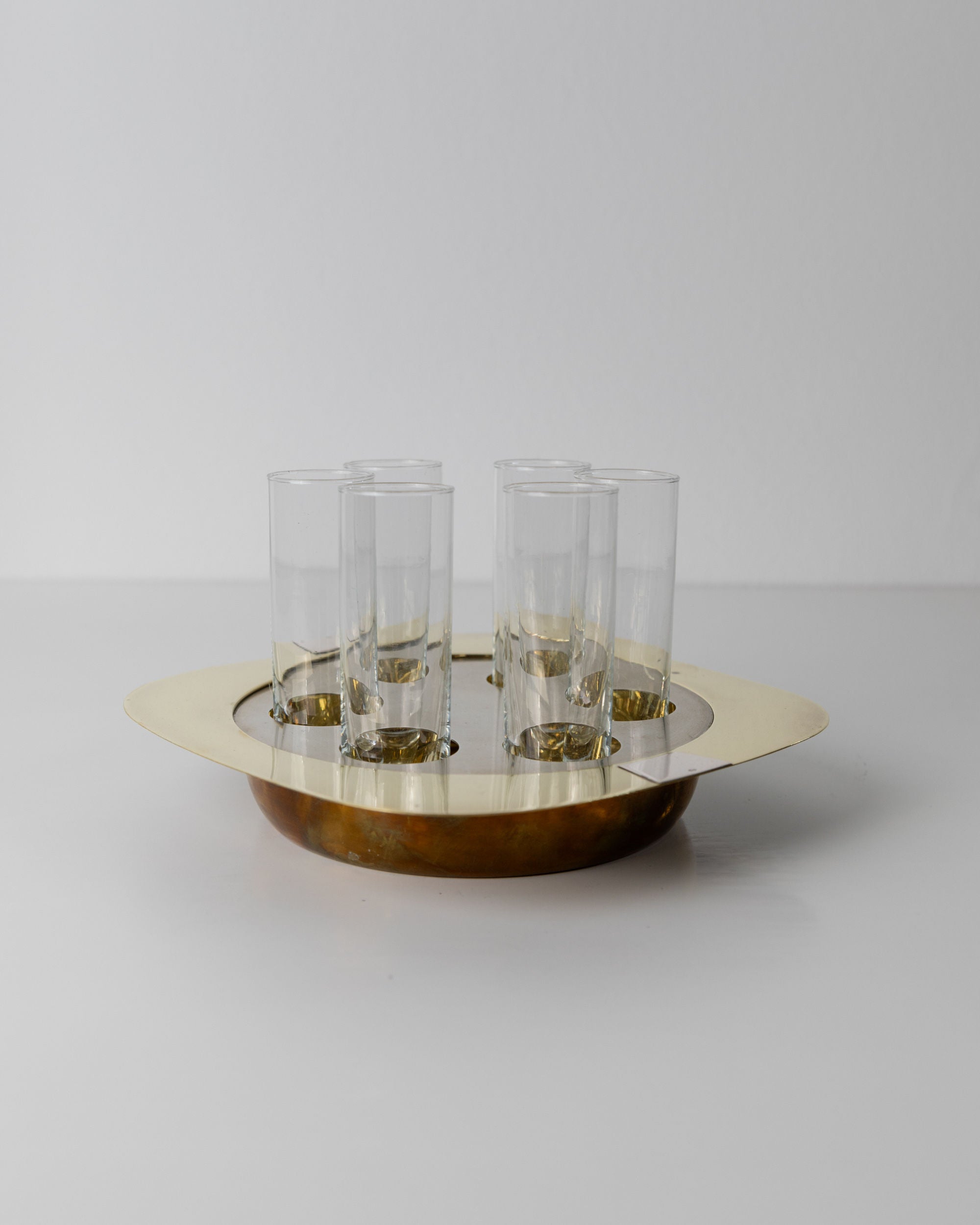 The Bottega Jacobs Set Shot Glasses and Tray features a round brass tray with six vintage shot glasses, each in its own holder, all resting on a white surface with a light gray background. Its shiny brass finish offers a reflective quality reminiscent of 70s mid-century design.