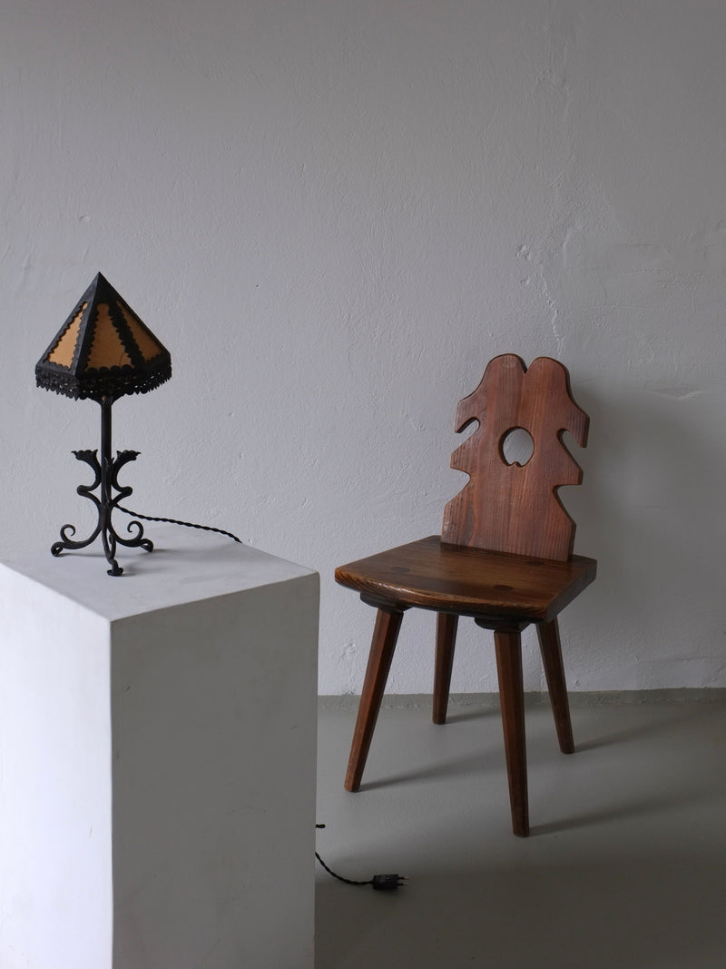 A wooden chair with a decorative backrest sits beside a white pedestal displaying the Veter Vintage Wrought Iron Dragon Table Lamp, Italy 1910s, with a triangular silk shade. The setup is against a plain gray wall.