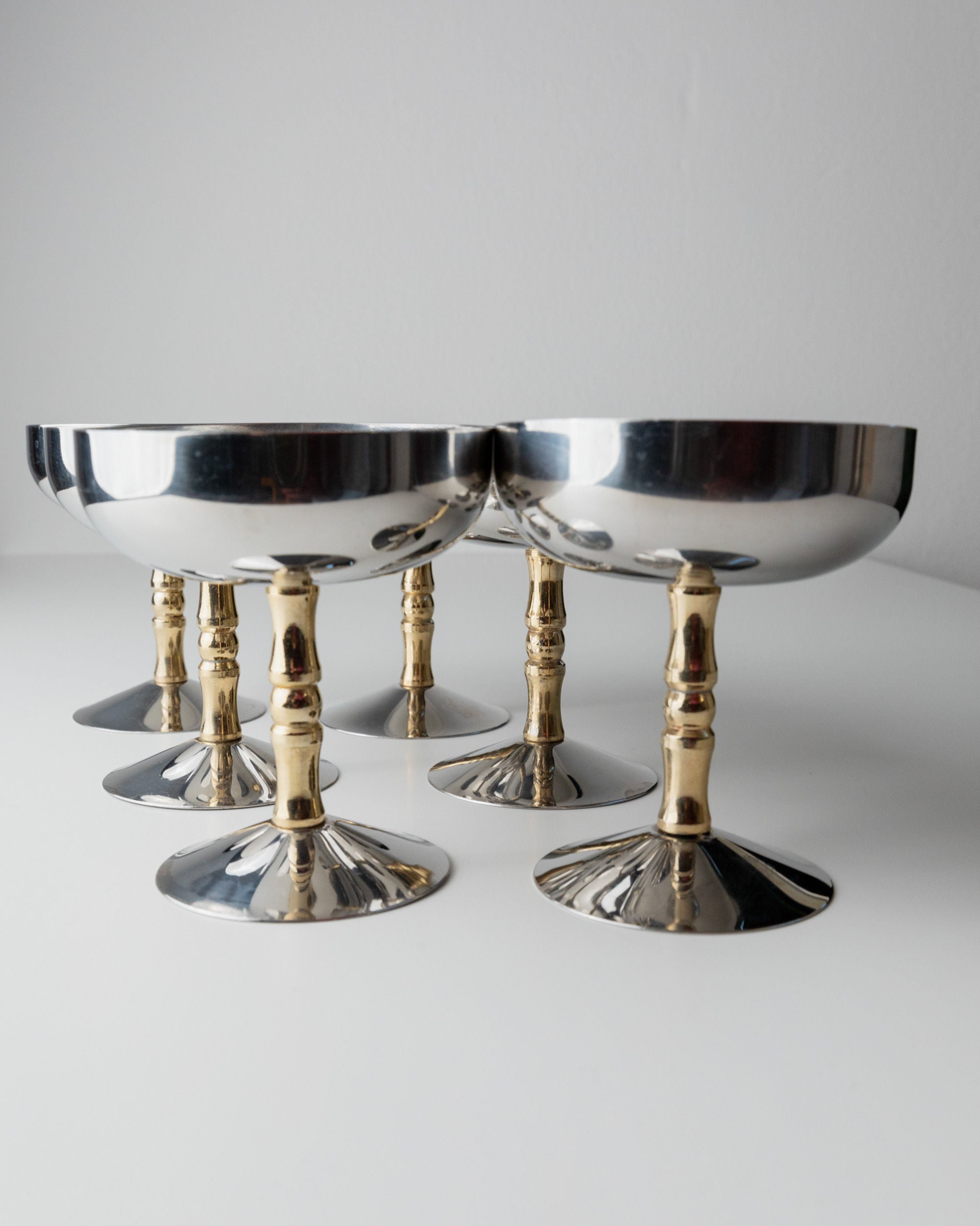 Six Bottega Jacobs 6 Stainless Steel Champagne Coupes 70s are set on a white surface, with their mirror-like finish contrasting the warm gold-toned stems, reflecting light and evoking timeless elegance.