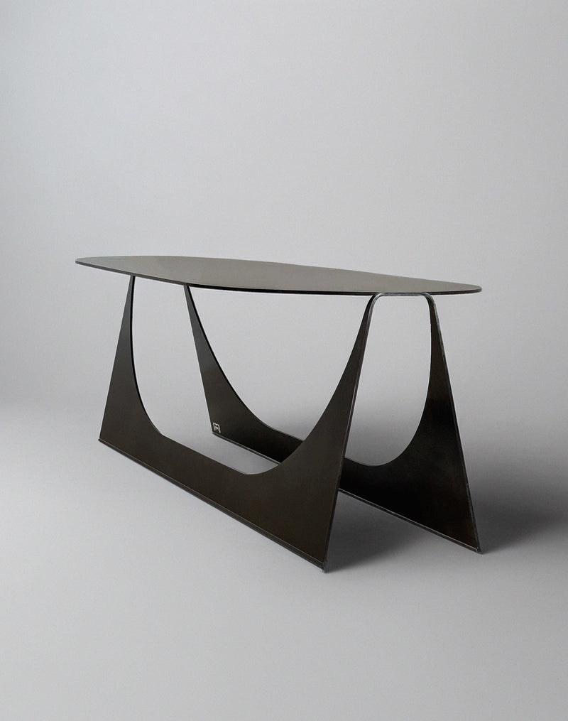 The 'Tani' Coffee Table by Modan is a modern, minimalist oval table in black steel with a slim top and geometric triangular base, shown against a plain light gray background.