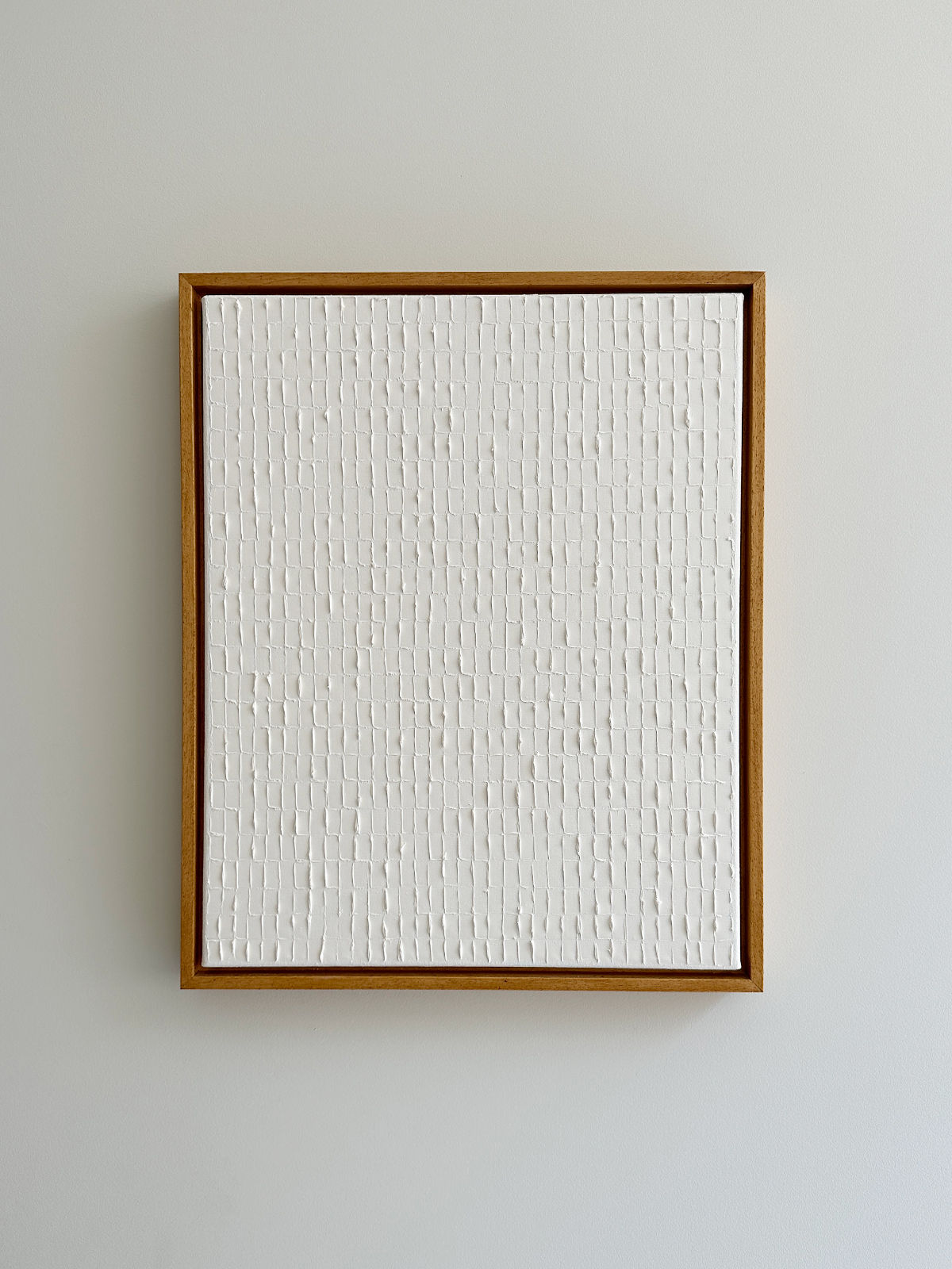 A rectangular artwork with a wooden frame features a textured white surface with a grid pattern, evocative of the Korean Dansaekhwa movement. The minimalist piece, Warm White - Oil on canvas by Roi Elmaliah, with evenly spaced vertical and horizontal lines creating small rectangular sections, exudes mindfulness and is mounted on a light-colored wall.