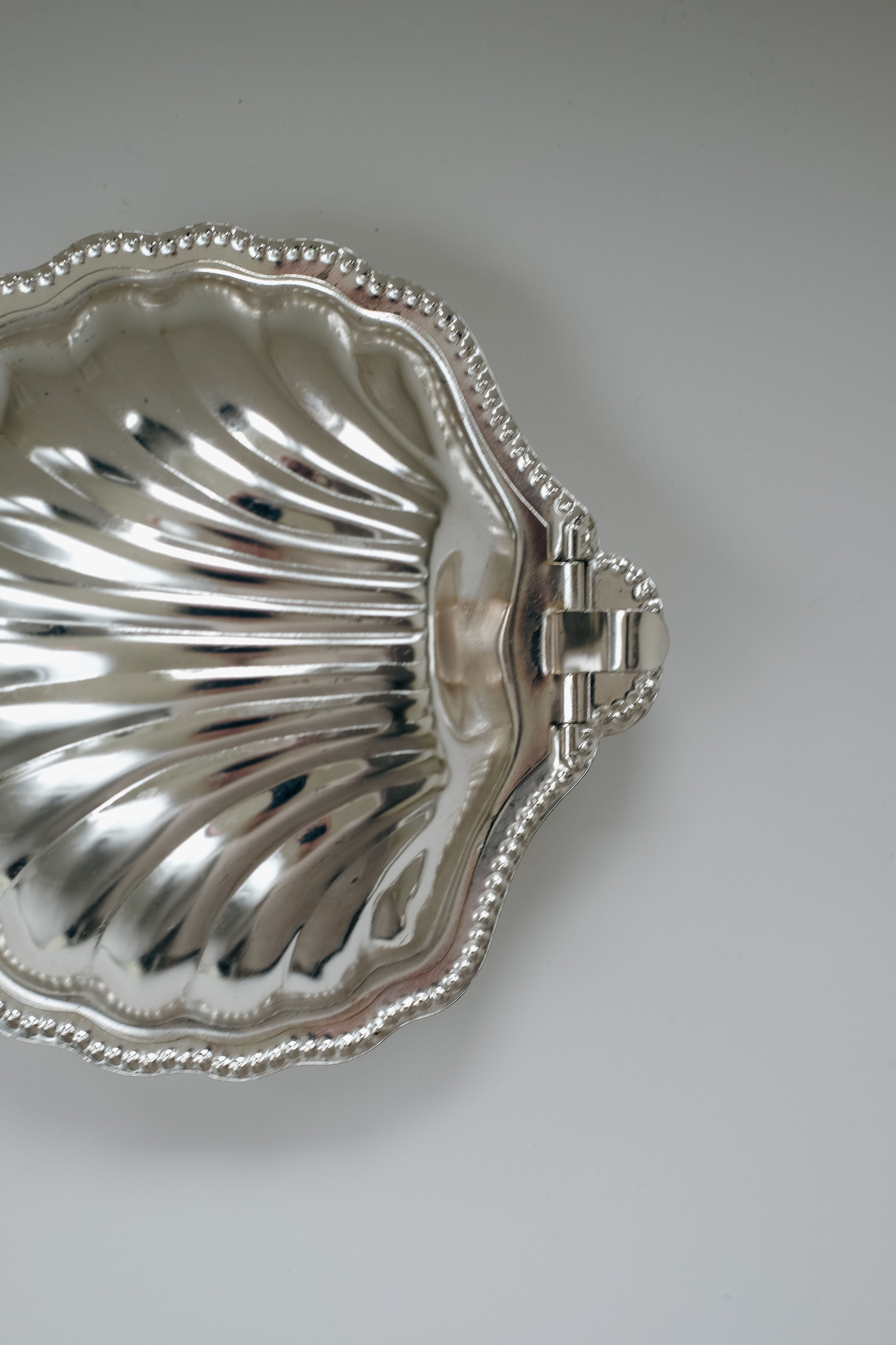 The Shell Shaped Butter Dish by septembre studios is silver with a seashell design, scalloped detailing, and right-side hinge. It features a beaded edge similar to vintage brass styles and is presented on a simple light background.