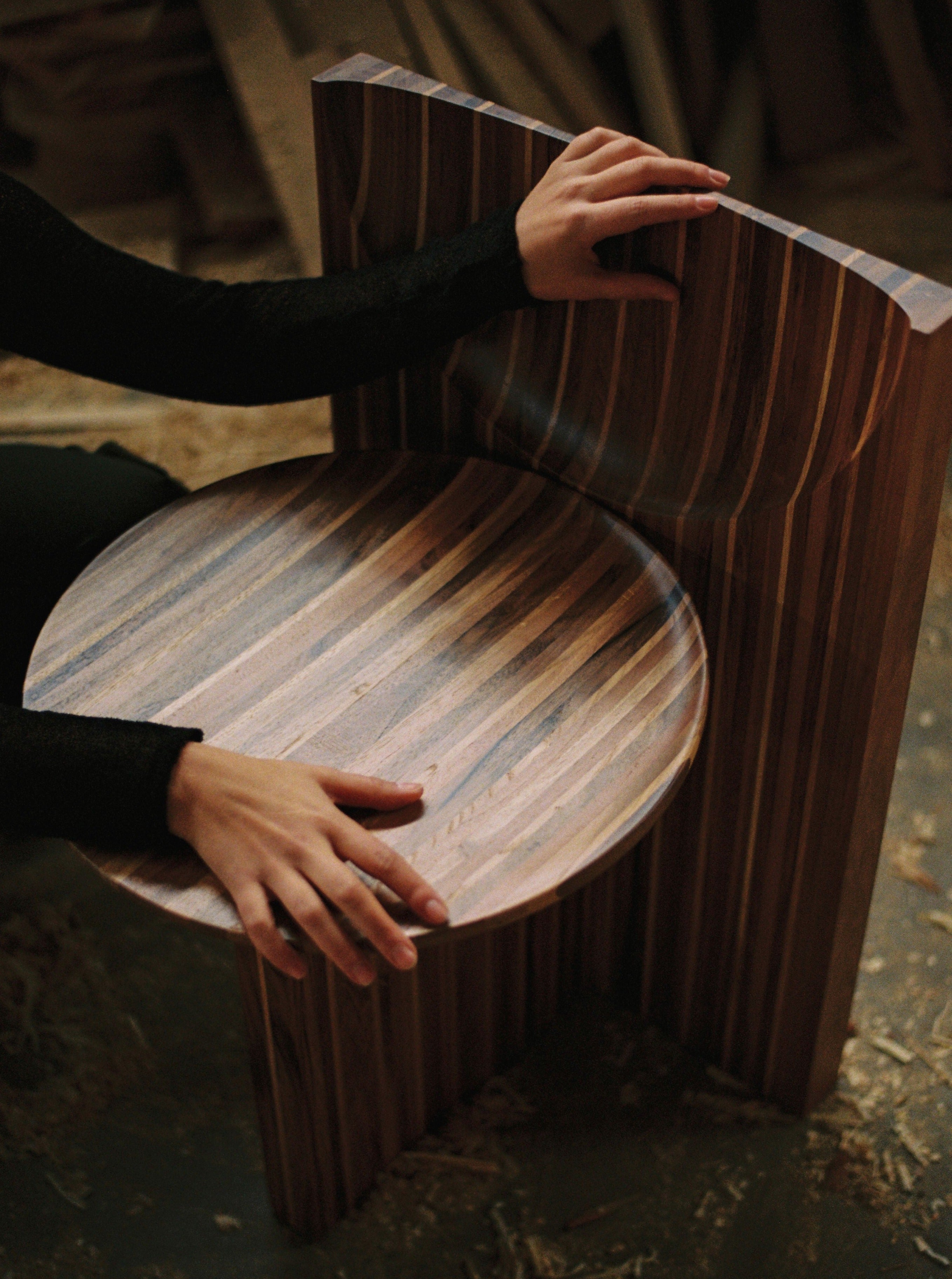 A person is assembling a Vaga Chair, a handcrafted piece with a unique design featuring vertical stripes of alternating colors. Made in Portugal from solid walnut and oak wood by the Rosana Sousa brand, the person's hands are seen positioning parts of the chair amid woodworking shavings on the ground, suggesting a woodworking workshop.