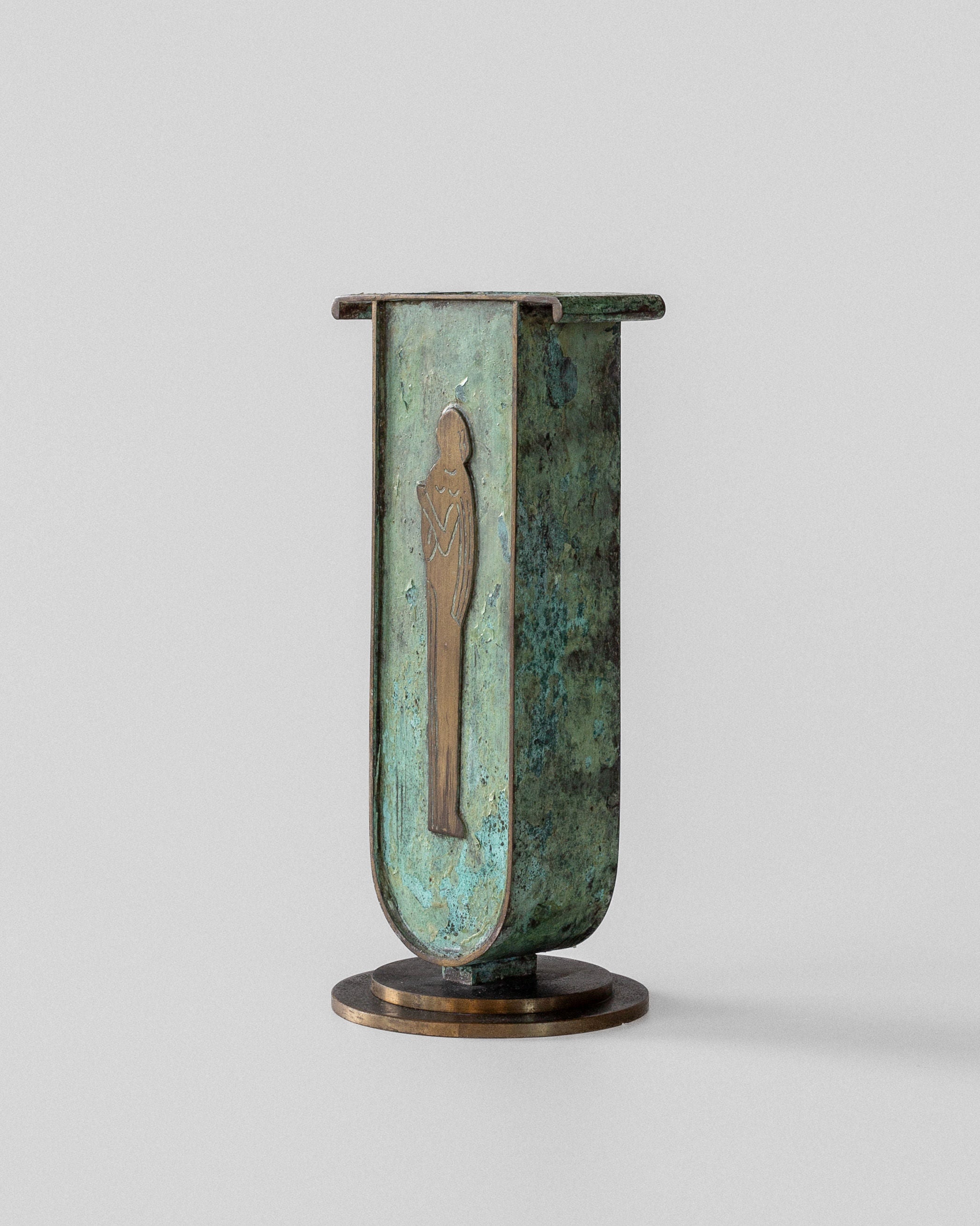 A Spigel Art Deco Brass Vase with a verdigris patina and engraved figure stands elegantly on a plain gray backdrop. Its rectangular form with rounded edges and circular base suggests Swedish influence, merging timeless elegance with modernism.