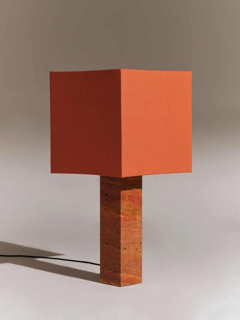 A Nassi Lamps Roma I Squared Red Travertine with an orange square fabric shade atop a matching square base. Crafted from natural materials, the lamp casts a soft shadow on a smooth, neutral-colored background. The design is modern and geometric, emphasizing clean lines and simple forms.