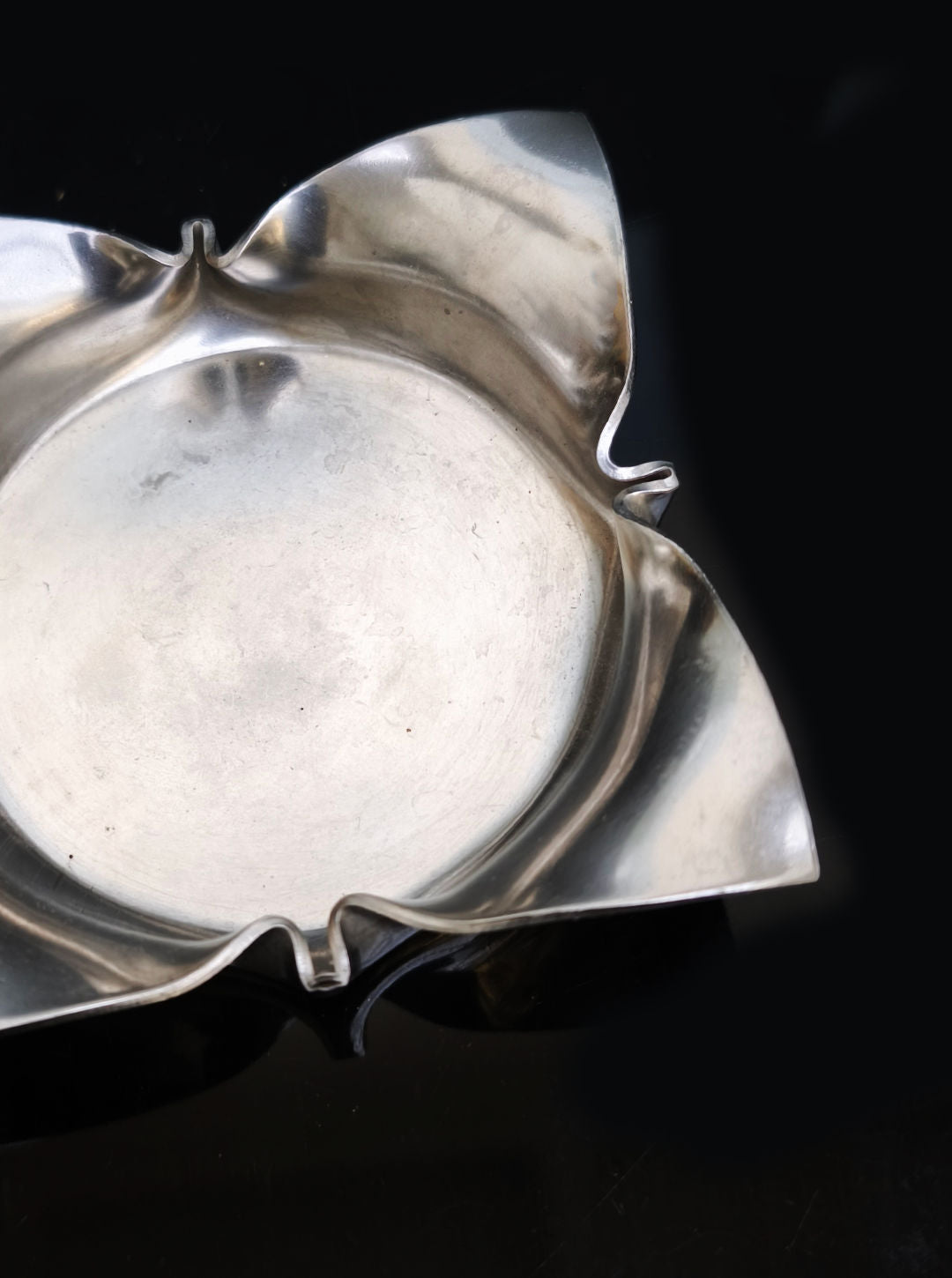 The Folded Ashtray by Les Objoies, a silver, flower-shaped brass piece with four petal-like extensions, rests on a glossy black surface. Its reflective metal and smooth design lend it a sleek, modern appearance.