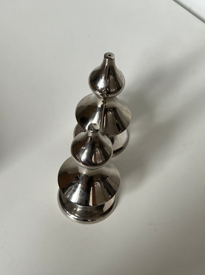 A silver-plated, chess-like piece in the shape of three stacked domes, each decreasing in size from bottom to top, is placed on a white surface. The Manushi Art Deco Salt & Pepper casts a soft shadow to the right.