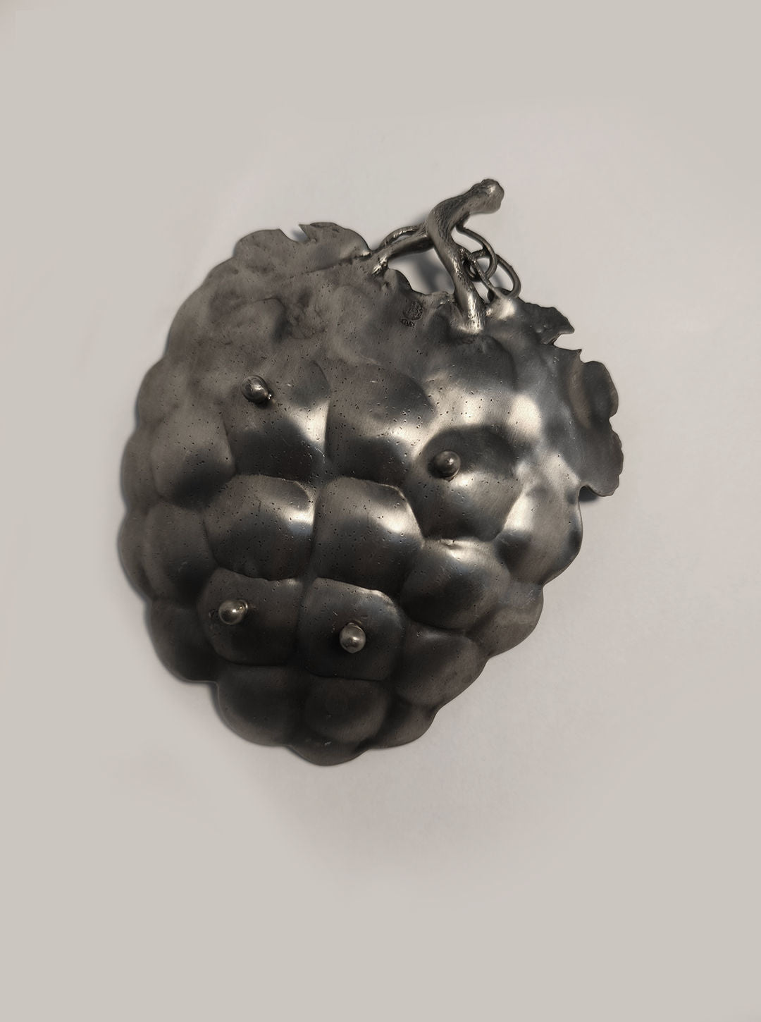 The Grape Vide Poches by Les Objoies features a textured metallic sculpture that resembles a cluster of raspberries with a small twig on top, making it an exquisite decorative piece. Its pewter-like elegance stands beautifully against the plain, light gray background.