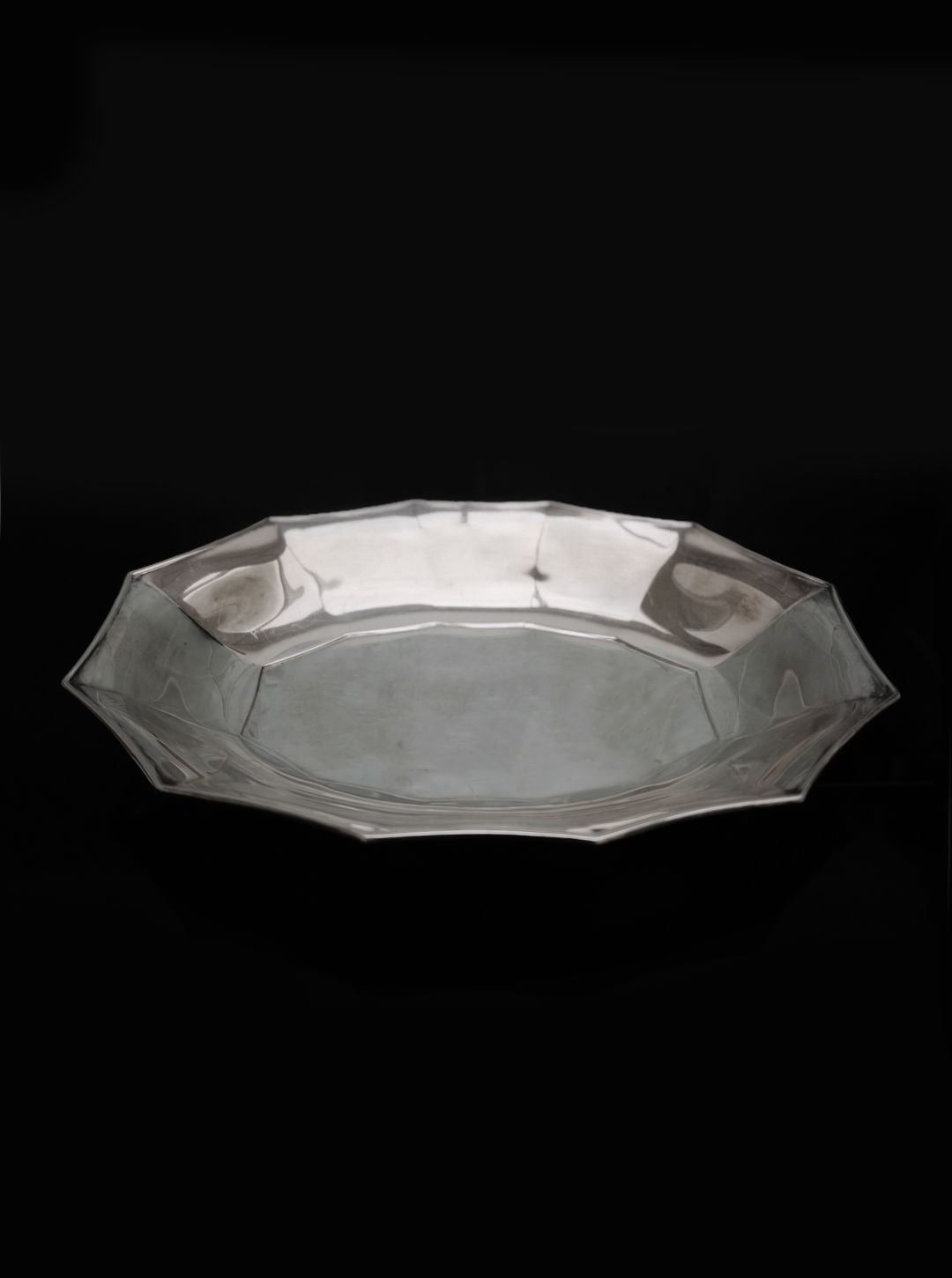 Set against a plain black background, the reflective Les Objoies Silver-plated Tray, featuring an octagonal shape with smooth surfaces and slightly raised edges, exudes sophistication.