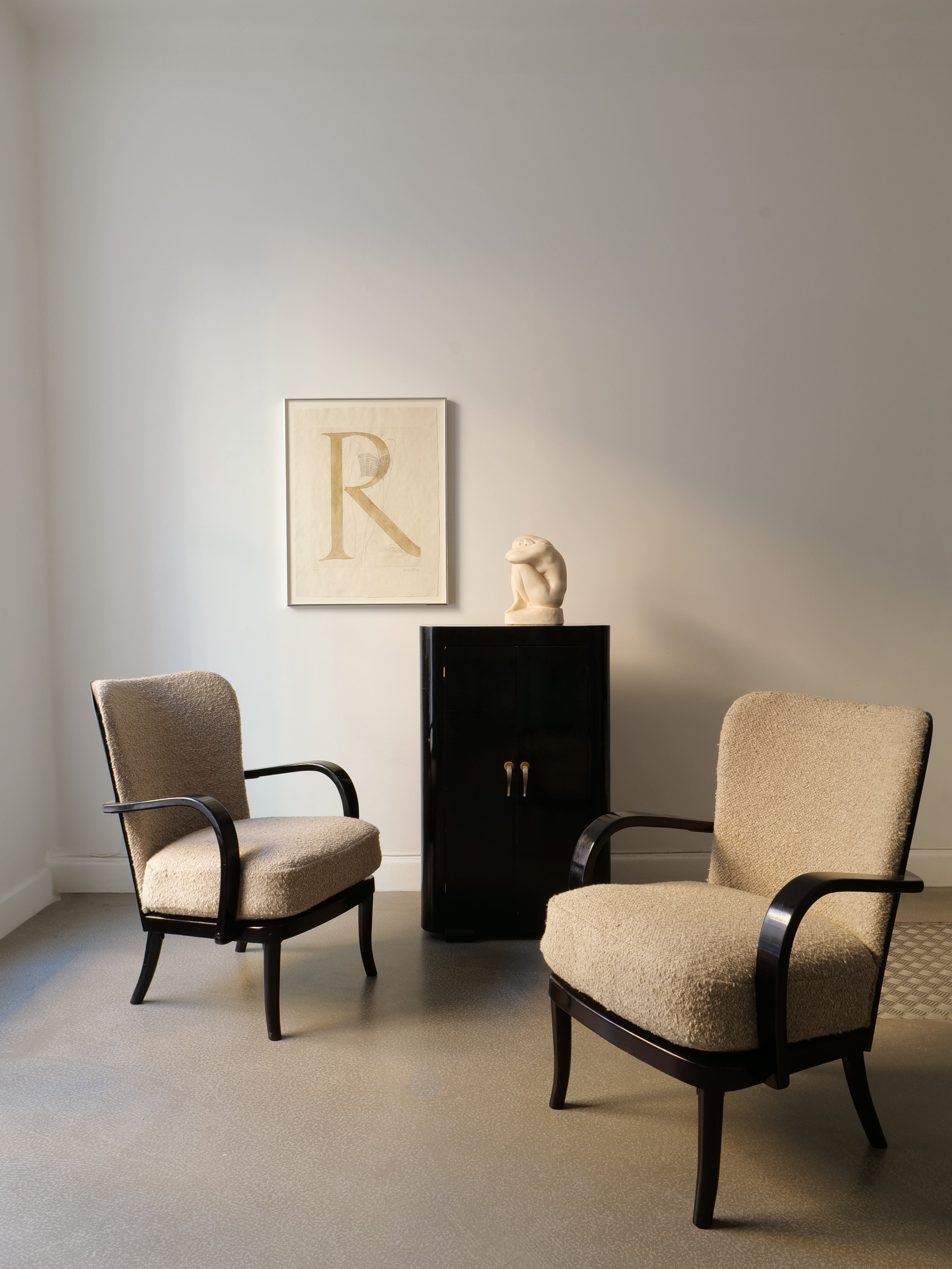 Two beige armchairs with dark wooden arms face each other in a minimalist room. A small black cabinet between them holds the Man Ray Etching 'Renee', 1970 from Collection apart. The light-colored wall behind is adorned with a framed letter "R," reminiscent of Man Ray's surrealist photography.