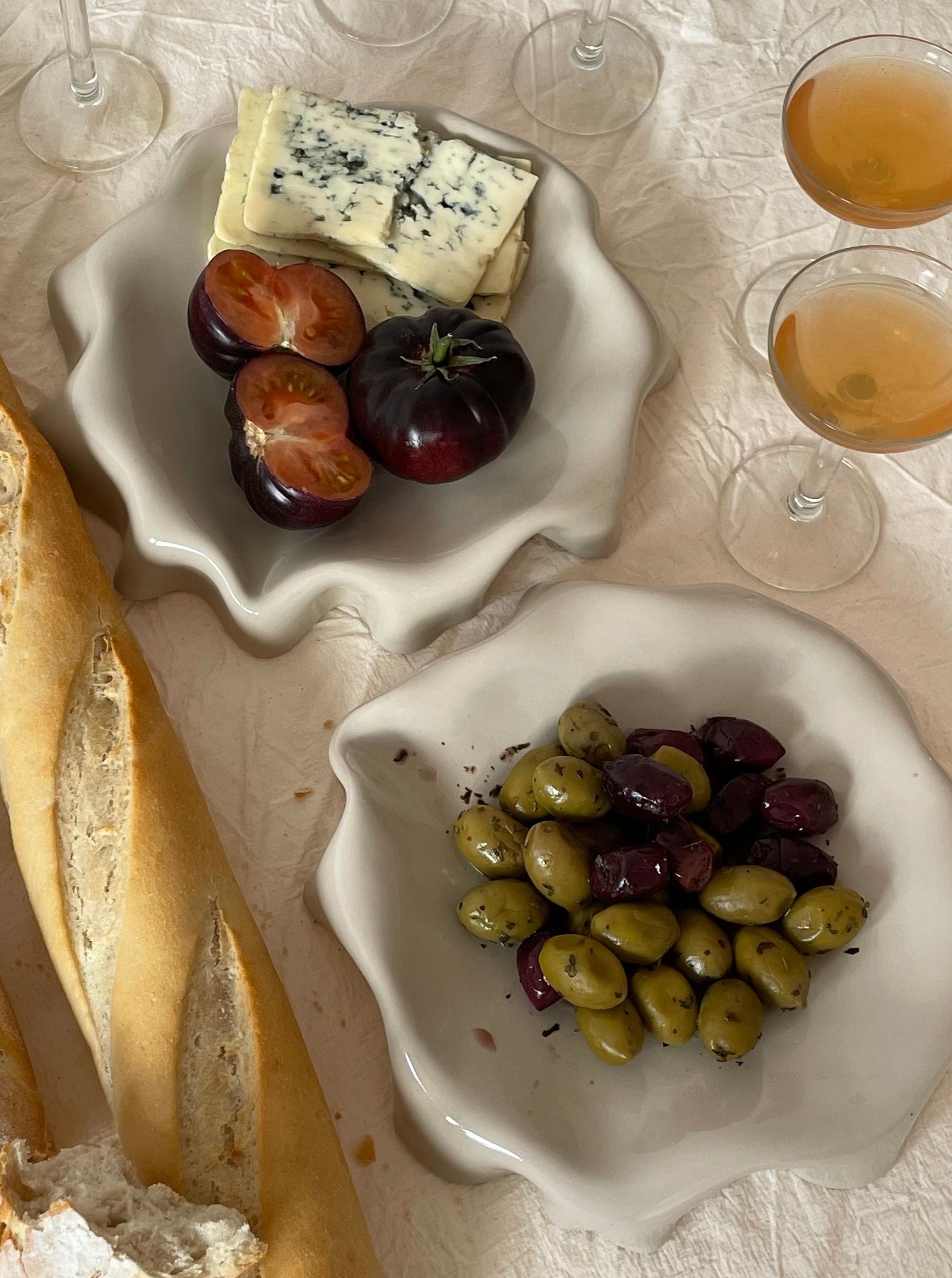 A table setup featuring a sliced baguette, two LESS Melted Butter Plates in Off White—one with assorted olives and the other with sliced tomatoes and blue cheese. Three glasses of a light-colored beverage are in the background. The scene is set on a white, textured tablecloth, showcasing practical tableware.