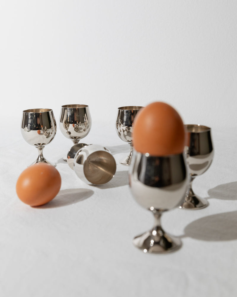 A set of six Bottega Jacobs egg cups from Italy, crafted in the 1980s, are elegantly arranged on a white surface. Two of these silver-plated egg cups cradle brown eggs, while one egg rests on its side. This arrangement exudes a minimalist and modern aesthetic, subtly reminiscent of mid-century design.