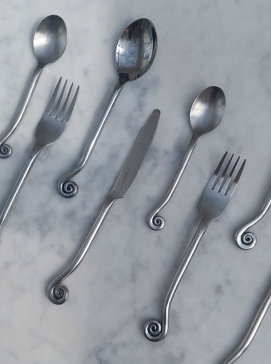 The Set of 4 Swirling Cutlery by Les Objoies features a stylish brass collection, including spoons, forks, and a knife with distinctive spiral-shaped handles, elegantly arranged on a marble surface.