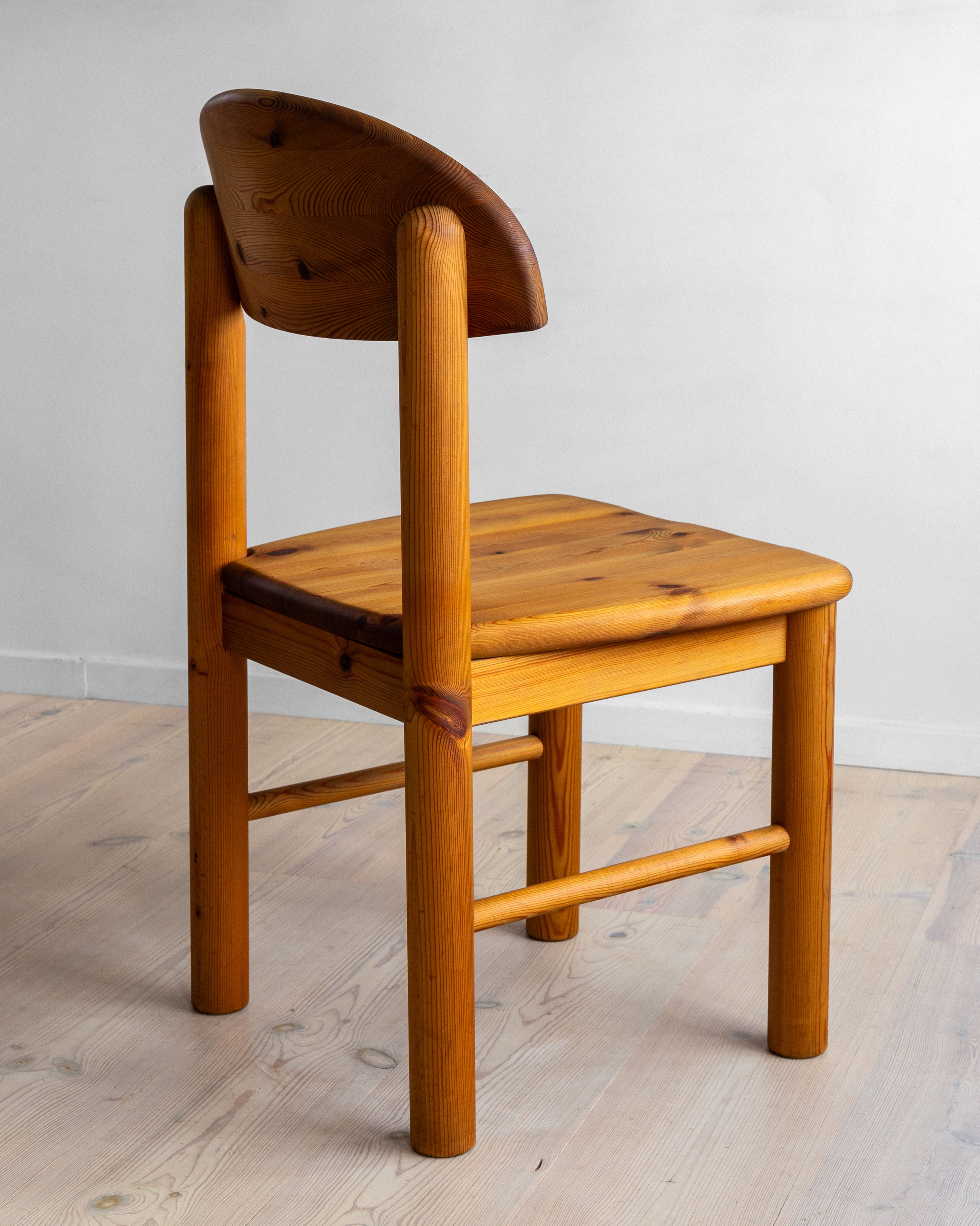 A set of four pine chairs by Rainer Daumiller from the 1970s, available from Spigel, are arranged on light wood flooring. These chairs boast a simple, rustic charm with curved backrests and sturdy seats made entirely from solid pine. Their thick, cylindrical legs are interconnected by horizontal supports, and they are set against a plain white background.