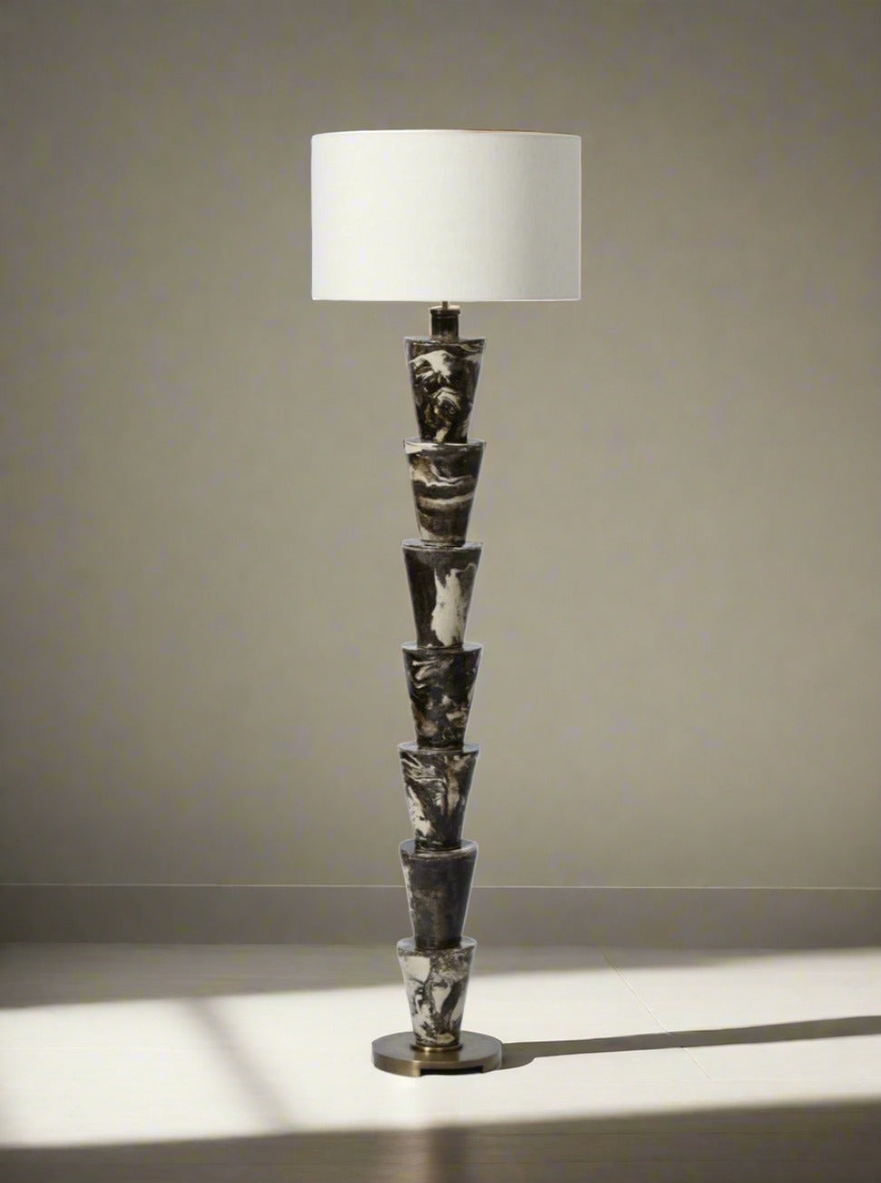 A unique "Nizwa" Floor Lamp by Barracuda Interiors with a series of stacked sculpted figures forming the base, topped with a white cylindrical lampshade. The marble-like base, featuring a predominantly black and white color scheme, stands on an iron base. This stoneware glazed floor lamp is exquisitely made in Portugal.