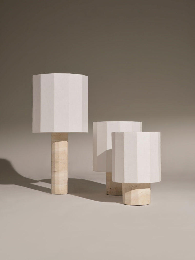 Three Roma II Dodecagon White Travertine lamps by Nassi Lamps with beige, cylindrical wooden bases and hexagonal white lampshades are arranged in a staggered formation in front of a plain background. The neutral tones and pure linen shades create a calm, modernist aesthetic.