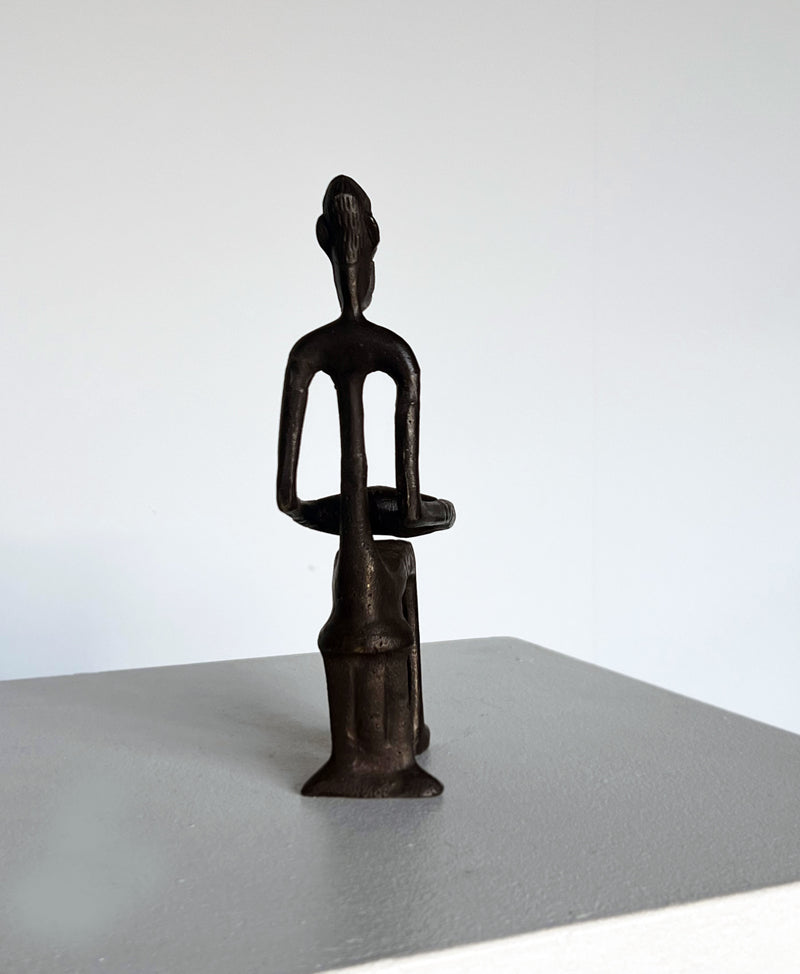 The Bronze Drummer Man Sculpture by Porter Studio is a dark bronze piece featuring a stylized human figure with elongated features, sitting on a small pedestal. The minimalistic details are viewed from the back, set against a light gray background on a plain surface.